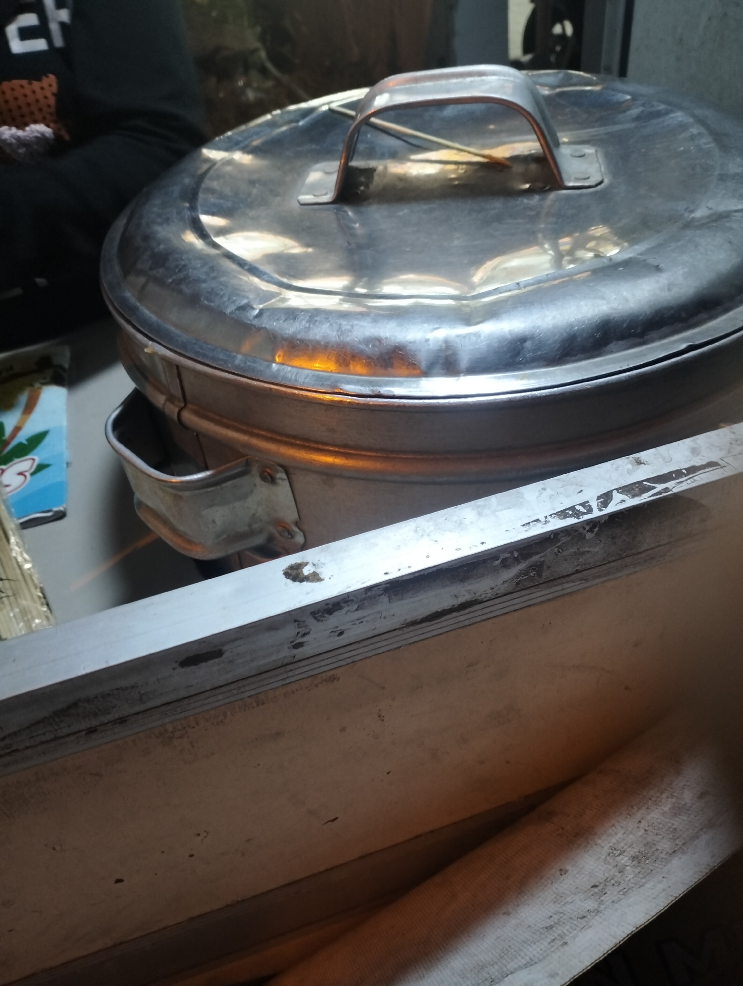 The image shows a close-up, slightly angled view of a large, round, metallic pot with a handle on its lid. The pot appears to be made of stainless steel and has a shiny, somewhat worn surface. There is an orange glow emanating from under the lid, suggesting the pot is heated or contains something hot. A person wearing a dark hooded garment is partially visible on the left side of the frame, with the back of their shoulder and arm in view. The person's clothing has a patterned section on the chest. In the background, there is some greenery and other indistinct objects. In the foreground, there is a textured surface, possibly a table or counter, with a metallic edge running horizontally across the bottom of the image. The overall lighting suggests an indoor environment, possibly a kitchen or food stall. The location is specified as Krikilan, Indonesia.