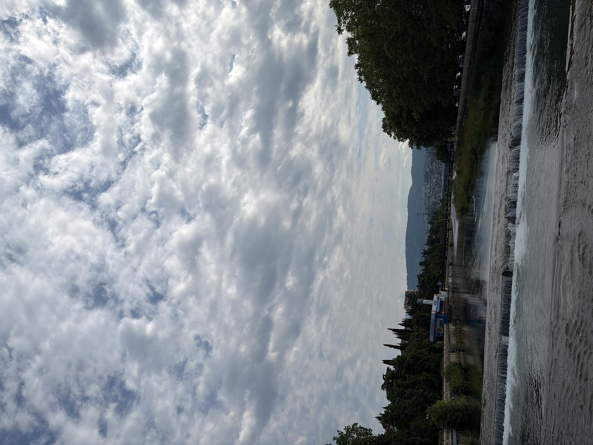 The image shows a view of the Mzymta River in Sochi, Russia, looking downstream towards the Black Sea. The river flows through a city with buildings and trees on either side. In the foreground, there is a small waterfall or dam in the river, creating white water rapids. The sky is cloudy with patches of blue sky visible. The overall scene is peaceful and natural.