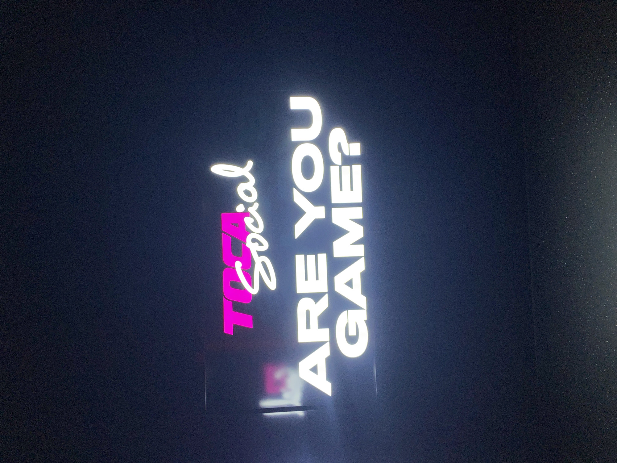 The image shows a vertical neon sign with the text "TOCA Social ARE YOU GAME?". The word "TOCA" is in a vibrant pink color, while "Social," "ARE YOU," and "GAME?" are illuminated in white. The sign is set against a dark background, likely an evening or nighttime setting. The sign is most likely at the TOCA Social location in the city of London, United Kingdom.