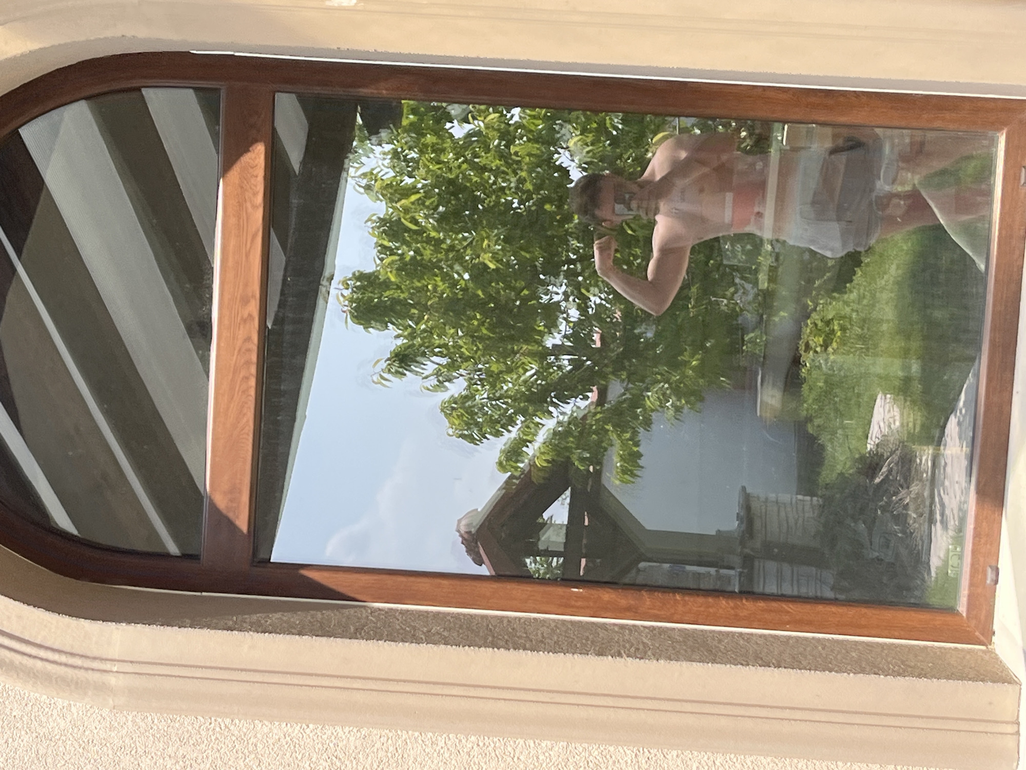 The image displays a wooden-framed window set into a textured, light-colored building exterior. The window features a rounded arch at its top and multiple glass panes.
Prominently reflected in the window is a person, likely male, seen from the chest up. They appear to be shirtless or wearing a very light-colored top, along with light-colored shorts or underwear. The person is posing, with their left arm flexed and elbow bent, seemingly looking towards the camera or their own reflection.
Beyond the reflection, and visible through the glass, is a vibrant green tree with abundant foliage, suggesting a lush outdoor environment. A structure with a pitched roof and a stone-like base is also visible in the background, along with green grass and other vegetation. The bright light and clear blue sky visible in the reflection indicate a sunny day.
On the interior side of the window, visible through reflection on the far left, are diagonal stripes, possibly from blinds or curtains. The building's exterior wall has a light, possibly stucco, texture and includes decorative molding around the window frame. There is no visible text in the image. The scene captures a bright, daytime moment, possibly a casual photo or selfie taken in Войводино, Bulgaria.
