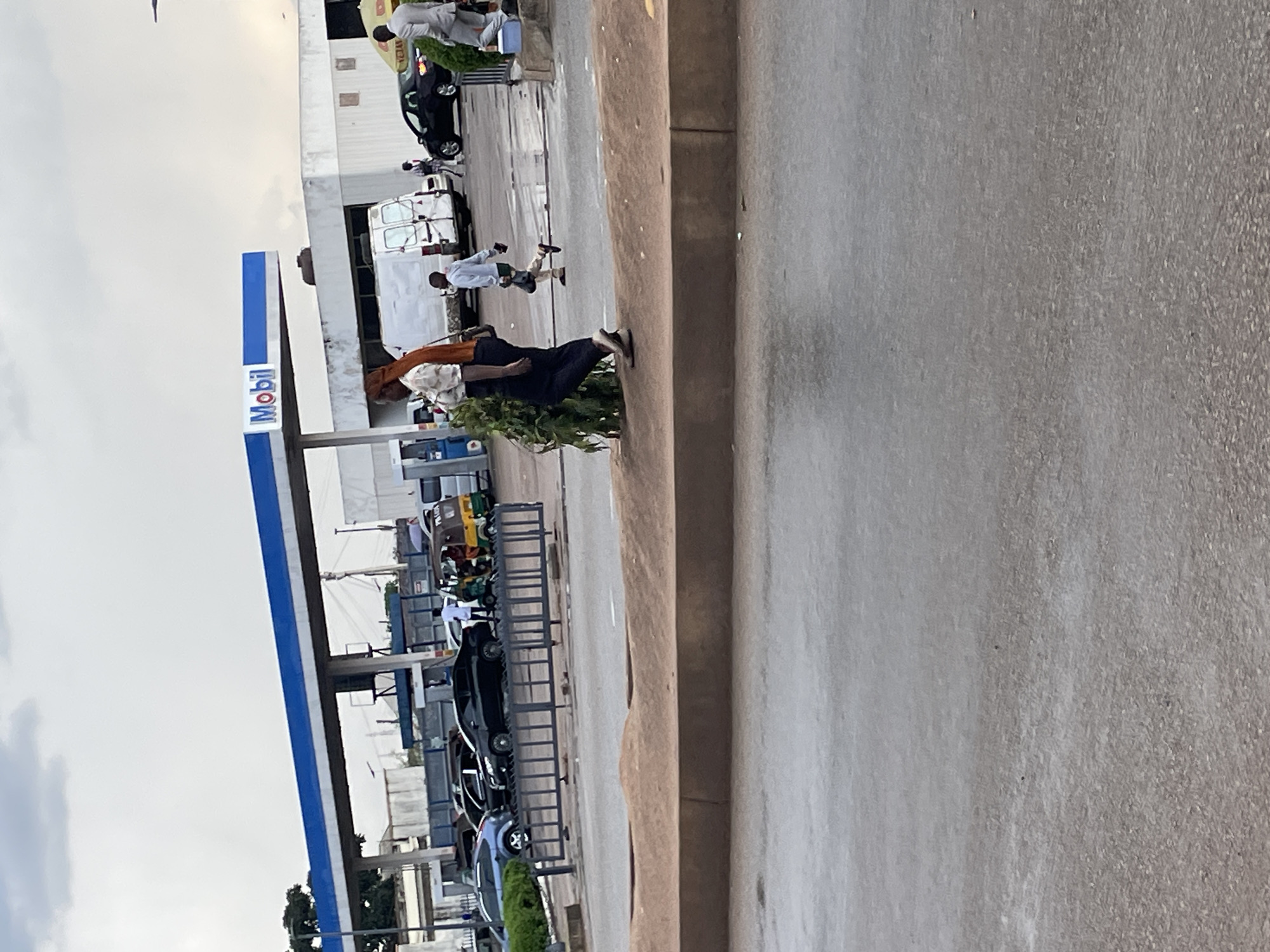 The image depicts a scene at a Mobil gas station in Jos, Nigeria. A gas station canopy with "Mobil" branding is visible. Several vehicles, including cars and a white van, are present at the station. A few individuals are seen walking around. In the foreground, a person in dark pants and a light-colored shirt with an orange head covering is walking past some foliage. Another person in a light gray hoodie and dark shorts is jogging. A tricycle, often used as a taxi, is parked. The ground appears wet, suggesting recent rain. The overall environment suggests an overcast day.