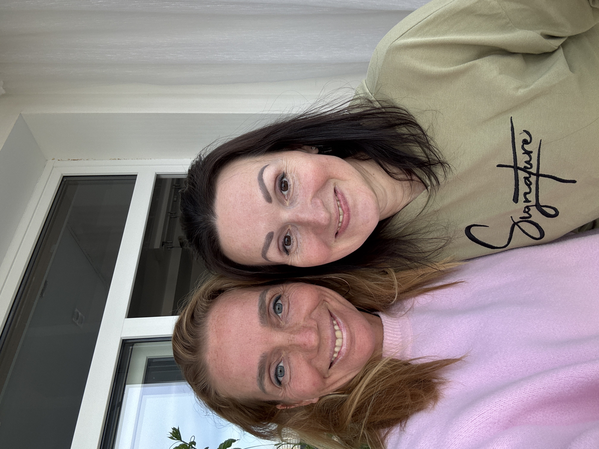 Two women are posing closely together. The woman on top has dark hair and is wearing a light green sweatshirt with a cursive "Signature" inscription. The woman below has blonde hair and is wearing a pink sweater. They are both smiling. The background includes a white window frame, possibly indicating an interior location. There is no explicit evidence in the image confirming the location is Saint Petersburg, Russia.
