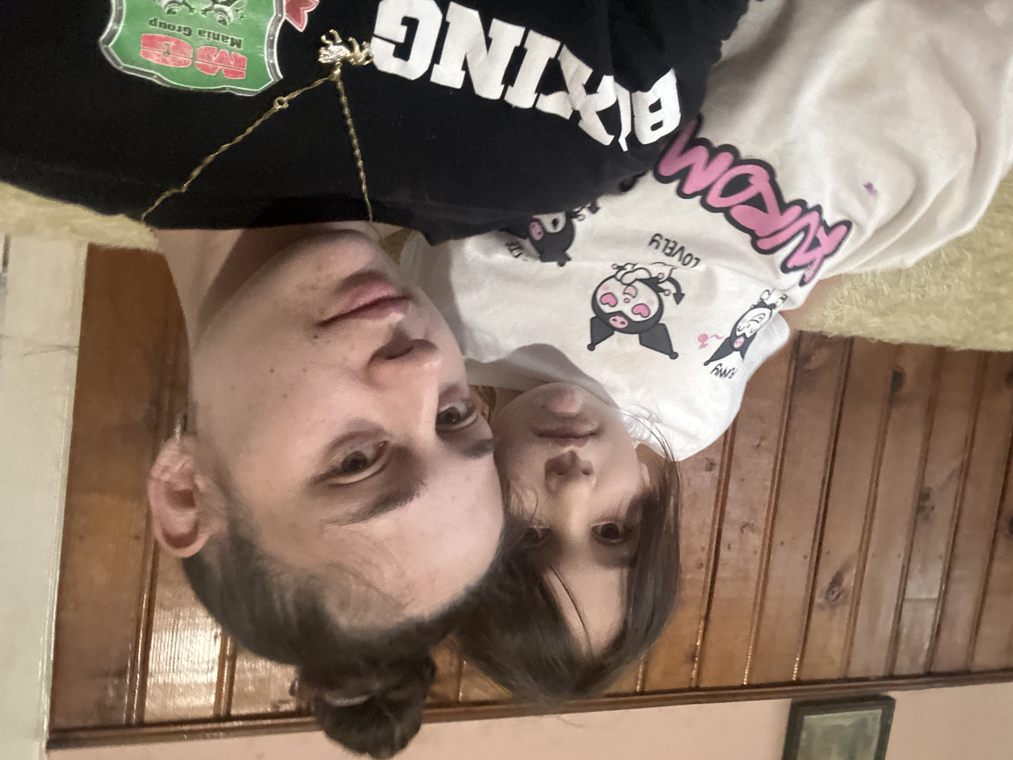 Two individuals are in the image. An adult and a child are closely positioned. The adult is wearing a black shirt with white text and a chain. The child is wearing a white shirt with cartoon characters. The background appears to be a room with wooden paneling. The image does not show any indications of the city of Resen, Bulgaria. The location cannot be confirmed.