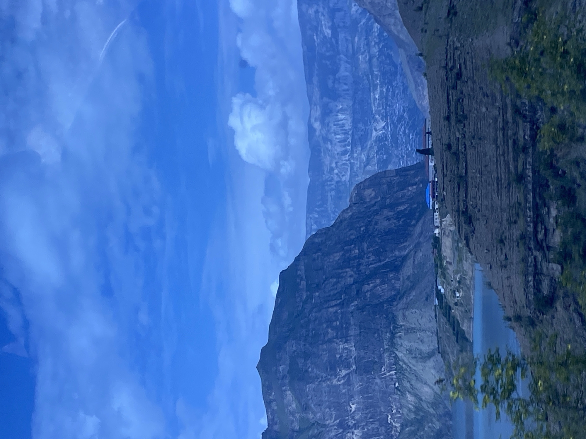 The image depicts a wide shot of a mountainous landscape with a body of water in the foreground. The sky is a deep blue with scattered clouds. A prominent, dark mountain range dominates the left side of the frame, with a lighter, more distant mountain range visible in the background. The terrain in the mid-ground appears to be terraced and arid, with several small buildings clustered on a hillside overlooking the water. The water itself is a vibrant blue. The overall impression is one of a remote and rugged natural setting. The location is Shamilkala, Russia.