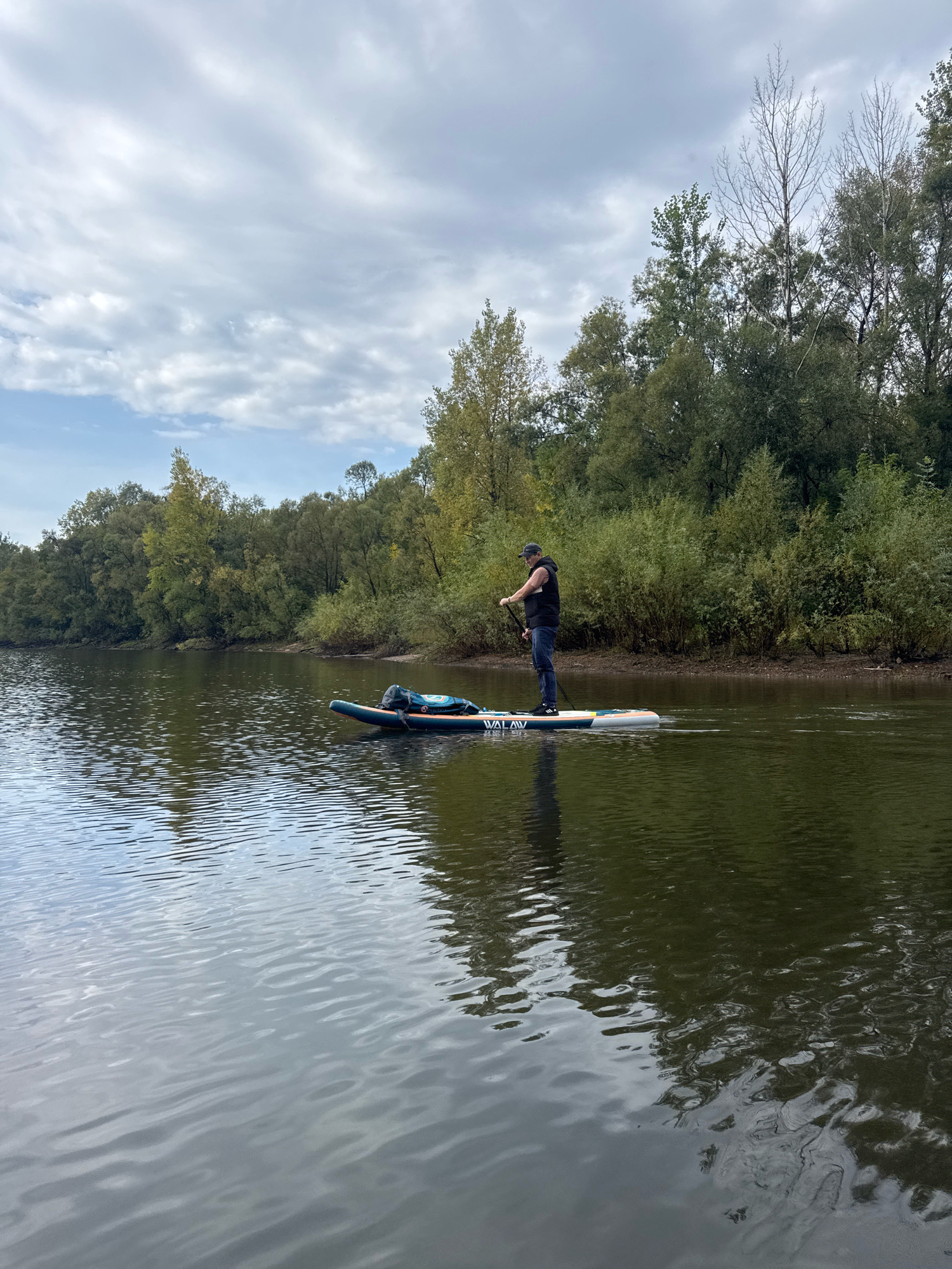 A person stands on a stand-up paddleboard (SUP) in a body of water, holding a paddle. The person is wearing a black vest over a dark shirt, blue jeans, and a baseball cap. The SUP is blue and white with orange accents and has a bag strapped to the front. The water is calm with ripples, reflecting the overcast sky and the surrounding trees. The background features a dense line of trees with green and yellow foliage, suggesting an autumn setting. The terrain appears to be a riverbank lined with vegetation. The image was taken in Ufa, Russia, along the Belaya River.