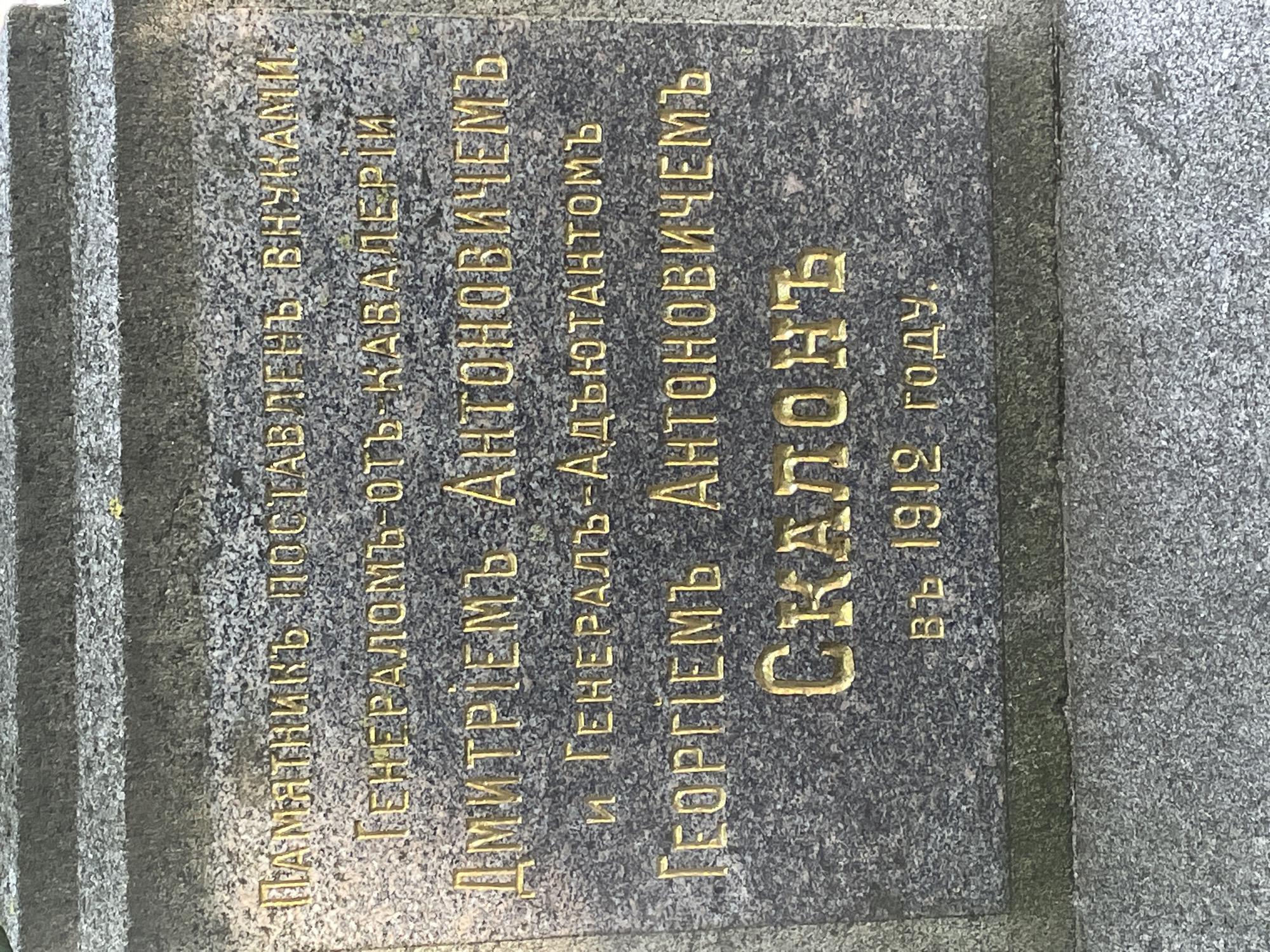 This is a close-up, eye-level, outdoor photograph of a granite monument with engraved gold lettering. The monument has horizontal carved lines at the top, suggesting it is part of a larger structure or a tiered memorial. The granite surface is speckled with gray and white, providing a textured background for the inscription. The gold lettering is worn in places, but largely legible.
The inscription reads:
ПАМЯТНИКЪ ПОСТАВЛЕНЪ ВНУКАМИ:
ГЕНЕРАЛОМЪ ОТЪ КАВАЛЕРИИ
ДМИТРІЕМЪ АНТОНОВИЧЕМЪ
И ГЕНЕРАЛЪ-АДЪЮТАНТОМЪ
ГЕОРГІЕМЪ АНТОНОВИЧЕМЪ
СКАЛОНЪ
ВЪ 1912 ГОДУ.
This translates to:
"Monument erected by grandsons:
General of Cavalry
Dmitrii Antonovich
and Aide-de-camp General
Georgii Antonovich
Skalon
In 1912."
The monument is positioned against a backdrop of more granite, with a distinct horizontal band of dark green, possibly moss or vegetation, running across the lower portion of the image, indicating it is situated outdoors, possibly in a park or cemetery. The lighting appears to be natural daylight, casting subtle shadows that highlight the depth of the engraving. There are no people or other subjects visible in the frame. The overall impression is one of historical commemoration.
The inscription and the style of the monument suggest this memorial is located in Russia, specifically in the city of Sof'ino, based on the provided context.