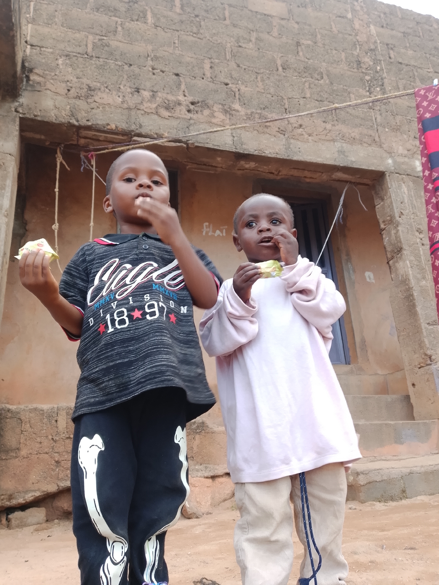 Two young children stand in front of a rough-hewn brick building. The child on the left, wearing a black t-shirt with white and red lettering and black pants with white bone graphics, holds a snack in their left hand and is bringing food to their mouth with their right. The child on the right, wearing a light pink long-sleeved shirt and light brown pants, is also bringing food to their mouth. The building has an open doorway and stairs leading up to it. A clothesline with a patterned fabric hangs across the upper portion of the scene. The ground in front of the building appears to be dirt or sand. The environment suggests an outdoor setting.