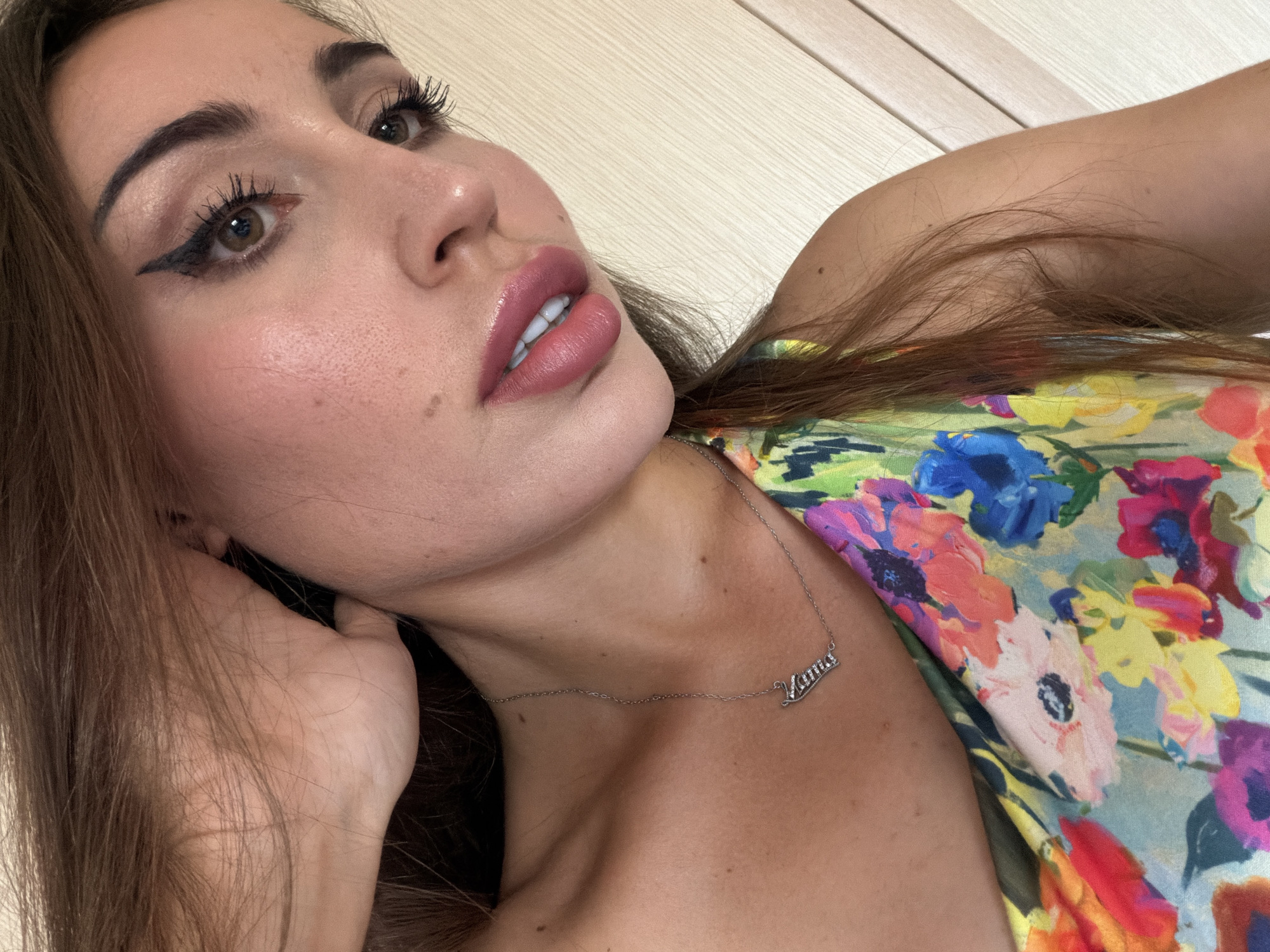 A close-up shot captures a woman's face from the chin up, angled to her right. She has long, brown hair that partially obscures her face and is touching her neck with her left hand. Her right eye is visible, with makeup including winged eyeliner and mascara. Her lips are parted, revealing her teeth. She is wearing a delicate silver necklace with a charm that appears to spell out a name. Her floral print top is also visible. The background consists of light-colored vertical panels, suggestive of an indoor setting. The image does not provide any information about a location in Tersefanou, Cyprus.