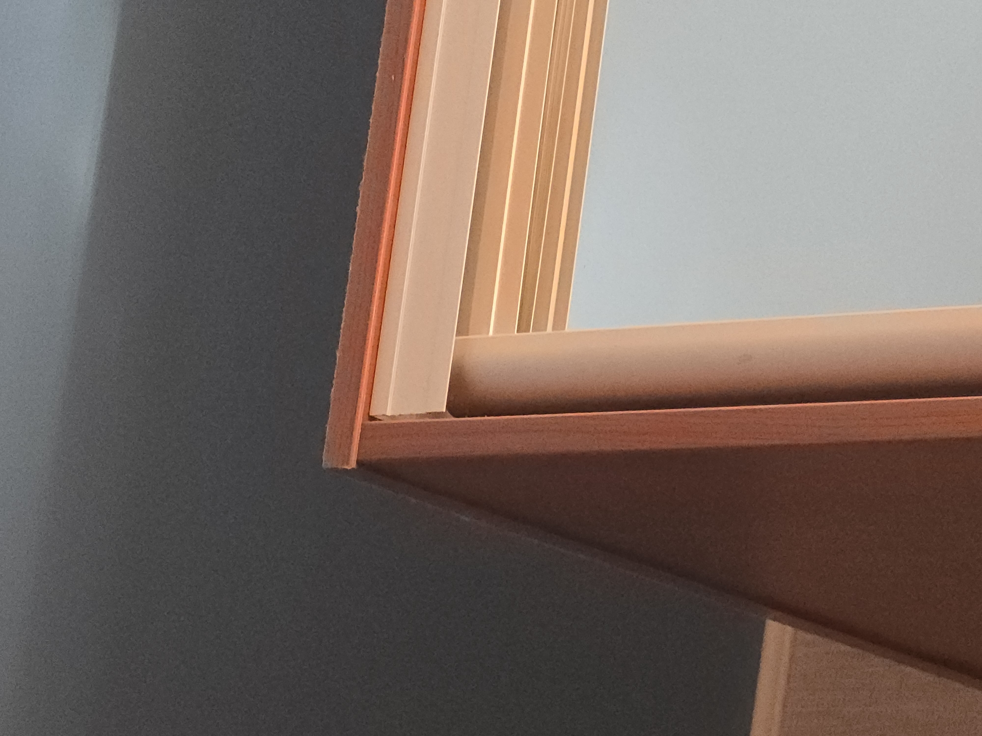The image is a close-up, abstract shot of interior architecture. The primary subjects are elements of a doorway or window frame, seen from an oblique angle. The frame itself is constructed of wood with a warm, reddish-brown tone, and appears to be polished or varnished. Surrounding the wooden frame is a lighter, possibly painted or laminate material, with a pale cream or beige color. This lighter material has multiple parallel, vertical indentations or grooves, creating a ribbed or fluted effect. There is also a thin, bright orange line visible, possibly a detail of the frame's construction or a contrasting insert.
The background on the left is a dark, solid color, appearing to be a wall that is partially in shadow, creating a dramatic contrast with the illuminated frame. On the right side of the frame, there is a smooth, light blue surface, which could be another wall, a ceiling, or the interior of a cabinet or room. The lighting suggests an indoor setting, with light coming from the right, casting a shadow onto the left wall. There is no clear indication of the time of day or weather, as the scene is entirely indoors and abstract. No people or animals are visible, and there is no text. The overall impression is one of clean lines, material textures, and contrasting colors and light.
