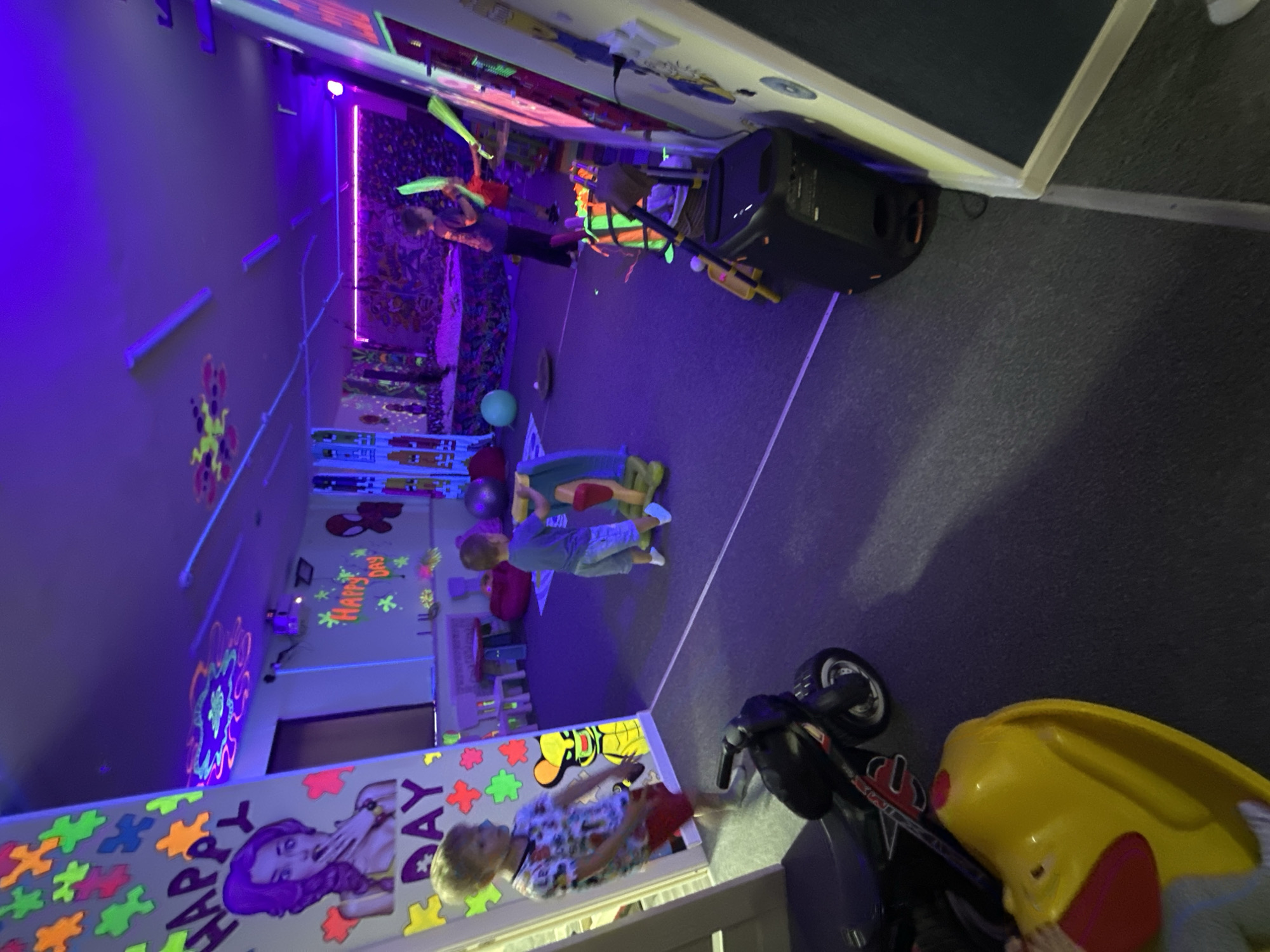 The image depicts a brightly lit room, predominantly illuminated by purple and neon lights. The setting appears to be a children's party or play area, possibly a birthday celebration, given the visible text that reads "HAPPY DAY". The walls are adorned with colorful, glowing decorations, including puzzle pieces and abstract shapes, characteristic of a blacklight party theme.
In the foreground, a young boy with blonde hair, wearing a patterned shirt and red shorts, is standing. To his right, a black ride-on toy, possibly a motorcycle, is parked next to a yellow plastic toy with red accents. In the background, two children are interacting. One child, wearing a blue shirt and jeans, is sitting on the floor, while another child in a green and orange striped top is standing or leaning. A large speaker or sound system is visible in the upper right portion of the image. Scattered throughout the room are various objects like a blue ball and other party decorations. The overall atmosphere suggests a fun and energetic event for children.