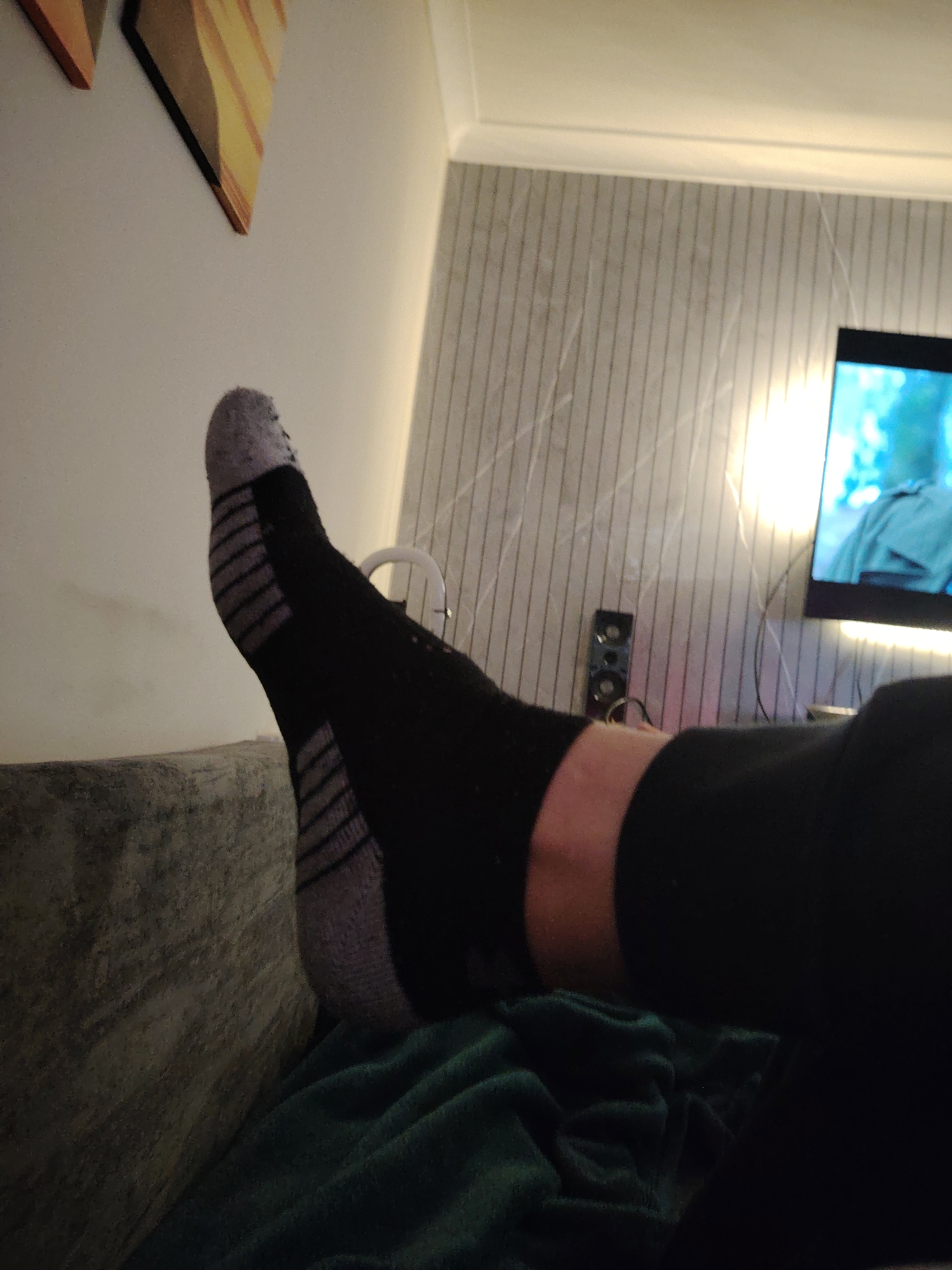 The scene depicts an indoor domestic environment, located in Huntingdonshire, United Kingdom. The foreground features a person's elevated left foot, clad in a black athletic sock with a grey toe and several prominent horizontal grey stripes on the sole, indicating grip features. The ankle skin is visible and light-toned, while the leg is covered by dark fabric. The foot rests near a textured, grey upholstered surface, likely part of a sofa or bed. Below the foot, a crumpled dark green blanket is visible. The background consists of a light-colored wall with vertical striped paneling and a bright, vertical strip of light on the right. Two abstract art pieces with geometric patterns in orange, yellow, and brown tones are mounted on the upper left wall. On the right, a flat-screen television displays a blue-toned image, showing a figure in a light blue garment. A small black electronic device with two visible circular components sits below the television, with associated cables. The ceiling, featuring decorative molding, is visible at the top of the frame. The ambient lighting is dim, suggesting a relaxed, evening setting.
