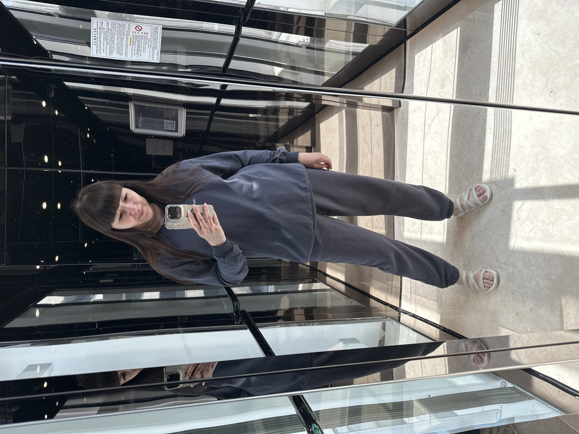 A woman is standing in an elevator, taking a selfie in the mirrored interior. She is wearing a dark blue sweatsuit and white sandals. The elevator interior is modern with black metal framing and mirrored walls. Signs and labels are visible within the elevator car. The elevator floor is light-colored tile. The reflection of the woman and elevator is seen in the mirrors. The location is Sochi, Russia.
