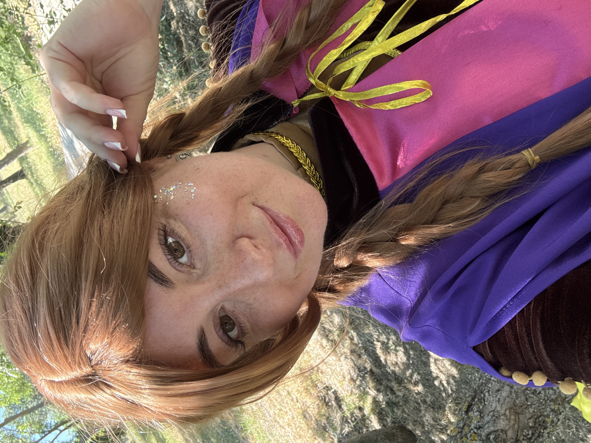 A close-up selfie of a young woman with auburn hair styled in two braids is taken outdoors during the day. She has freckles across her nose and cheeks and glitter applied to her left cheekbone. She is wearing a costume with a dark brown velvet top with gold embellishments, a purple cape draped over her shoulders, and a pink and purple dress with yellow ribbons tied at the chest. Her right hand is raised to her hair, and her fingernails are painted with a white and pink French manicure. The background shows a sunlit, grassy area with trees, suggesting a park or natural setting. The lighting indicates it is a sunny day, casting shadows on the ground.