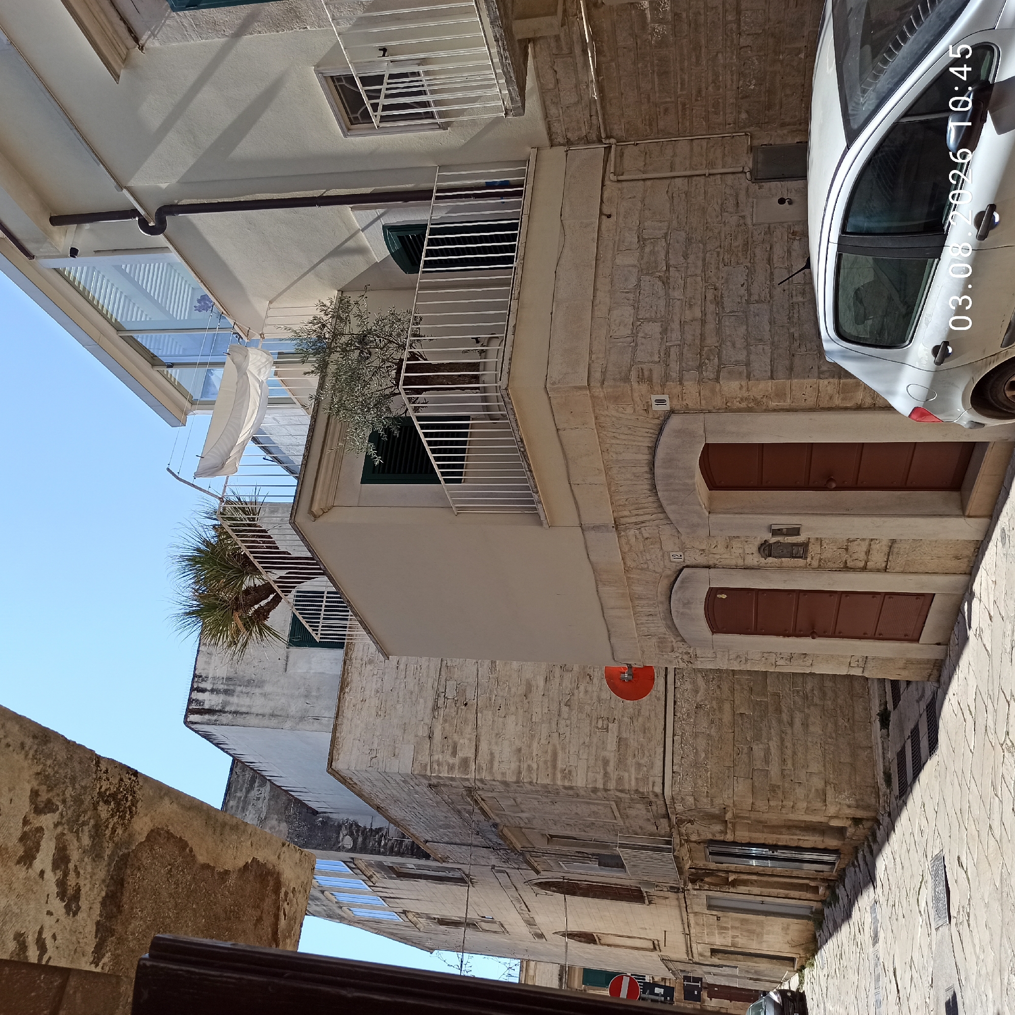 The image shows a sun-drenched, narrow street in the historic center of Ruvo di Puglia, Italy. The street is paved with irregular stone blocks, with shadows cast by the buildings on either side. On the right, a light-colored, four-door hatchback car is parked, partially obscuring the lower portion of a cream-colored building. This building features two identical dark brown doors with horizontal paneling, numbered '12' and '10' above their respective arched entrances. The upper part of this building has a balcony with a white railing, adorned with a potted olive tree. To the left, a taller, older building constructed of rough-hewn stone blocks rises. It has several windows, some with green shutters, and balconies with white railings. A palm tree is visible on a terrace of this building. A white sheet is drying on a clothesline strung between two buildings. The sky is clear and blue. A red circular sign with a white horizontal bar is affixed to the stone building on the left, and another identical sign is visible further down the street. The image is dated "03.08.2026 10:45".