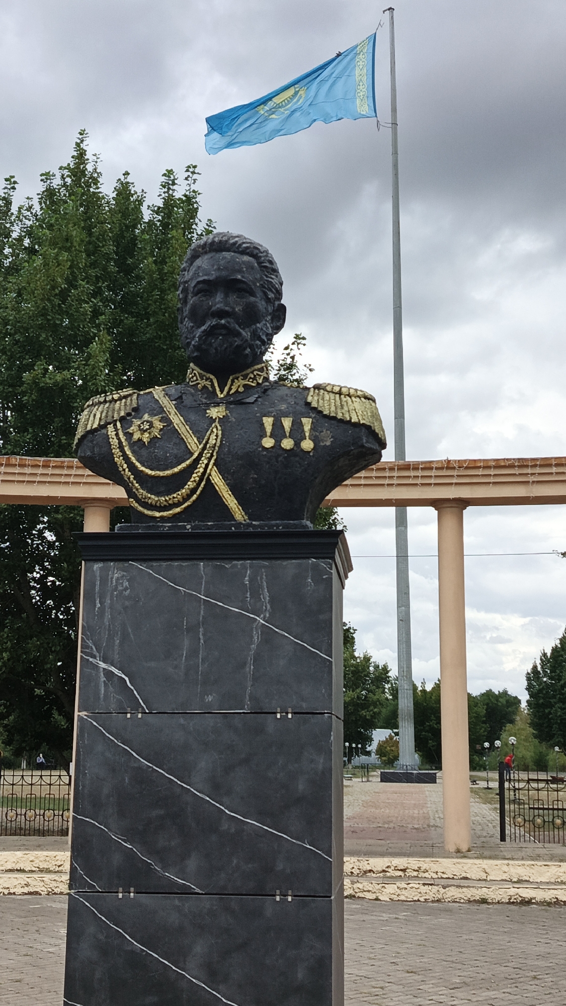 A dark, marble-like pedestal supports a dark bronze bust of a man with a beard, wearing a military uniform with gold epaulets and medals, and a sash. Behind the bust, a tall flagpole flies the blue and gold flag of Kazakhstan. The scene is outdoors, with trees and overcast skies in the background. The pedestal is made of dark grey rectangular panels with white veining. The bust is detailed, showing facial features and the ornate military attire. A beige structure with decorative lights runs horizontally behind the bust, supported by columns. A park-like setting is visible in the distance, with fences, pathways, and more trees.