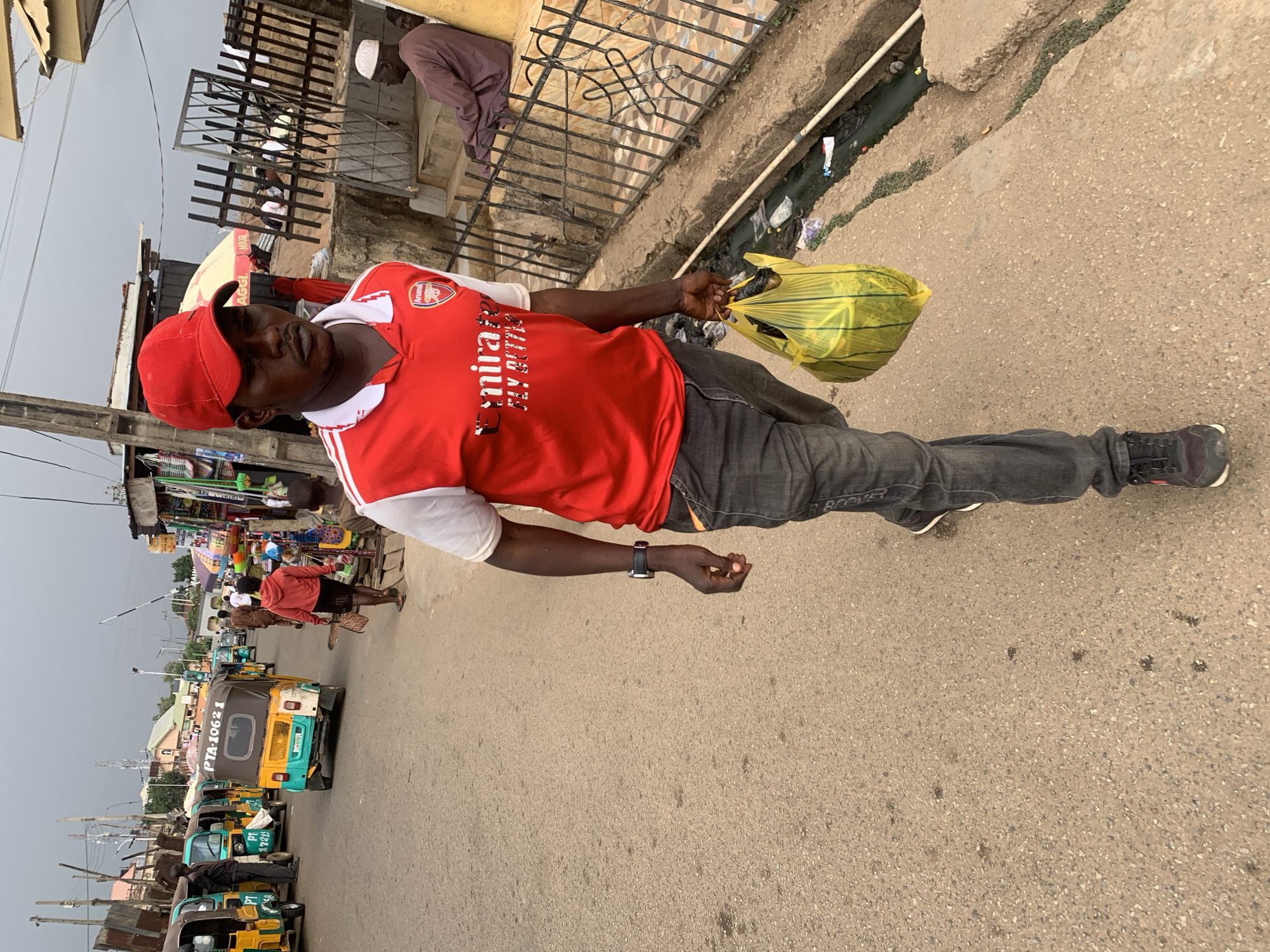 A man wearing a red Arsenal jersey and a red baseball cap walks down a paved street in Jos, Nigeria. He carries a yellow plastic bag in his right hand. He is wearing gray jeans and black shoes. A black wristwatch is visible on his left wrist. In the background are tricycles and people near storefronts, indicating a bustling urban environment. A gutter runs along the edge of the sidewalk, and a metal fence is on the other side. Another man wearing a white hat is standing by the fence.