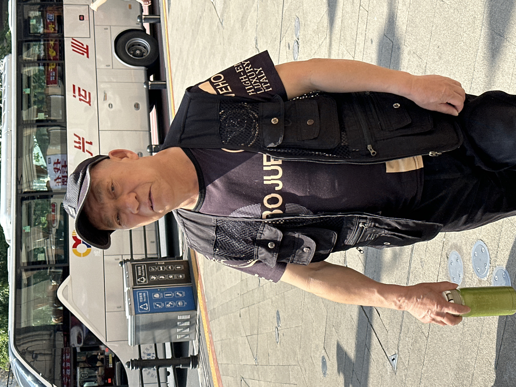 An eye-level, medium shot presents a man in Yuzhong District, China, holding a green container and looking at the camera. He is wearing a black t-shirt with the words "BO JUF" on it, along with a black utility vest that reads "VEHO HIGH-EN LUXURY ITALY" on the shoulder flap. A baseball cap covers his head, and he wears a ring on one of his fingers.
Behind the man, a white bus with red Chinese characters is partially visible. A trash and recycling receptacle is situated on the sidewalk. The ground is paved with square concrete slabs. The sunlight casts shadows across the scene.