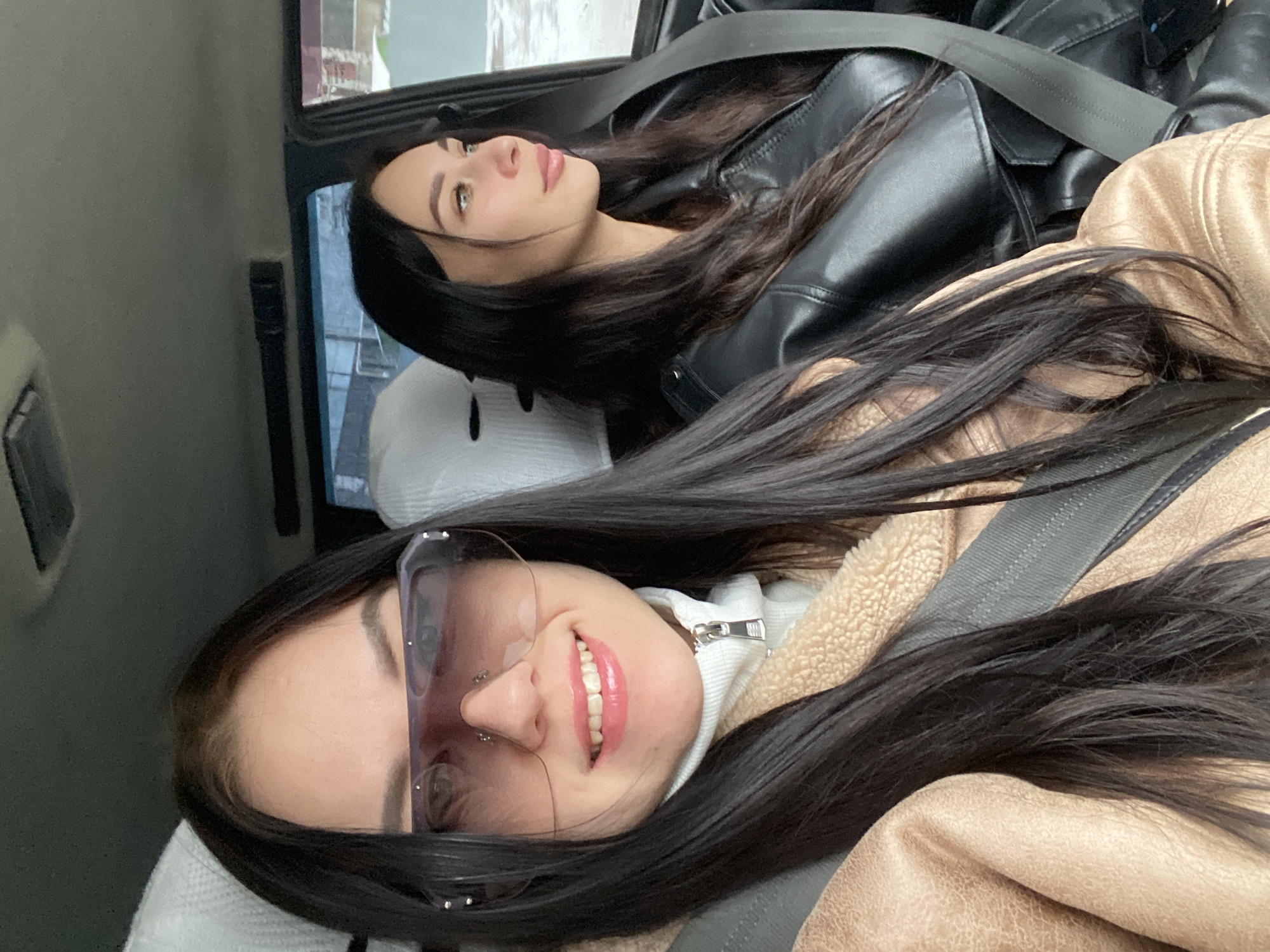 Two young women are reclined in the back seat of a car. The woman in the foreground wears sunglasses, a white turtleneck sweater, and a light brown jacket. She is smiling. The woman in the background wears a black leather jacket and is looking upwards. Both women have long, dark hair and are wearing seatbelts. Pillows are behind their heads. A partial view of a building is visible through the car window. The location is Chaikovsky, Russia.