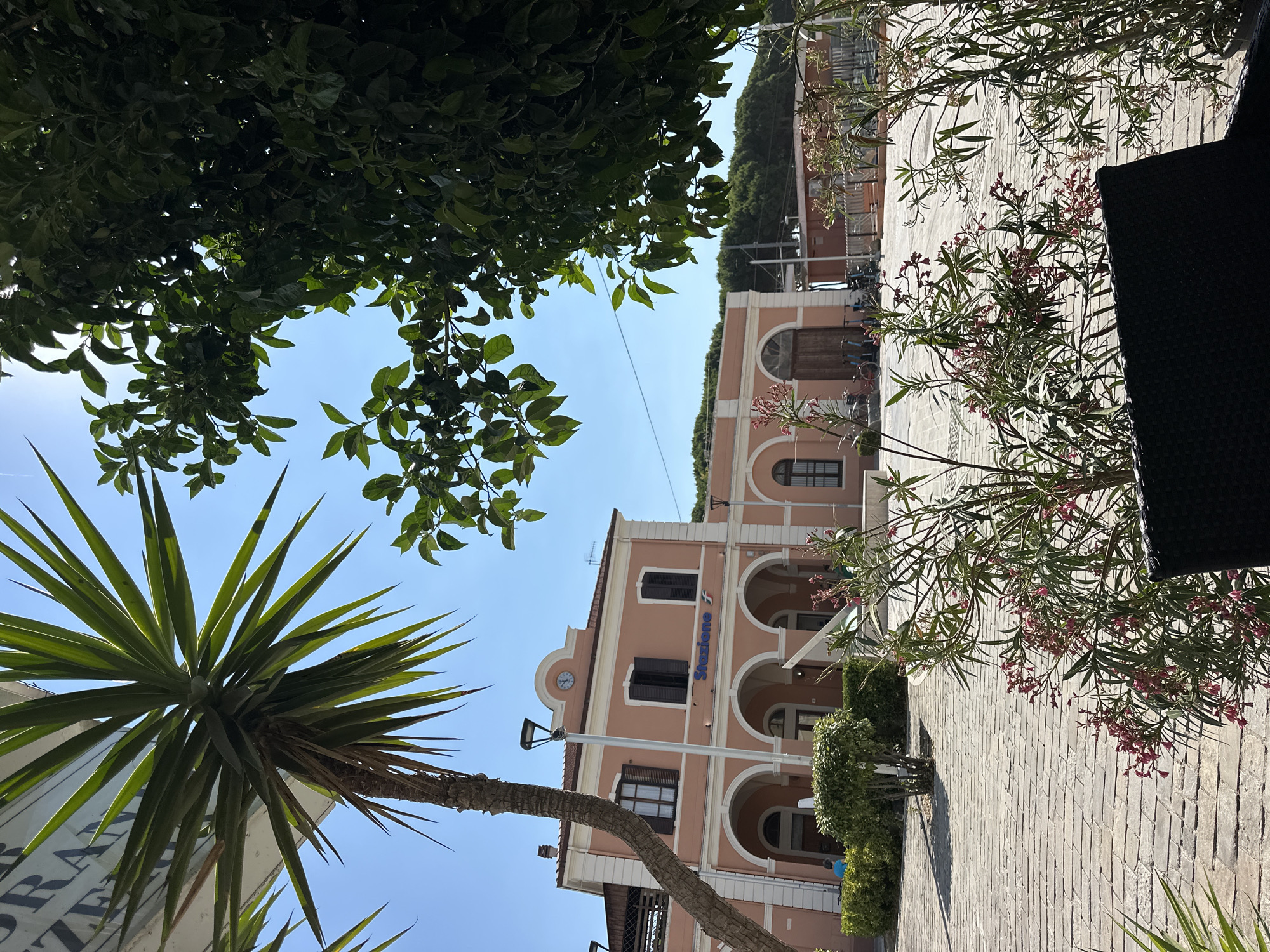 The image depicts a sunny outdoor scene in front of a train station in Pineto, Italy. A peach-colored building with arched entrances and windows is prominently featured, with "Stazione" and a train logo visible on its facade. A clock is mounted above the entrance. In the foreground, lush green trees and flowering bushes partially obscure the view of the station and the paved courtyard. A palm tree is visible to the left, and a black wicker chair is in the lower right corner. Several bicycles are parked near the station entrance. The sky is clear and blue.