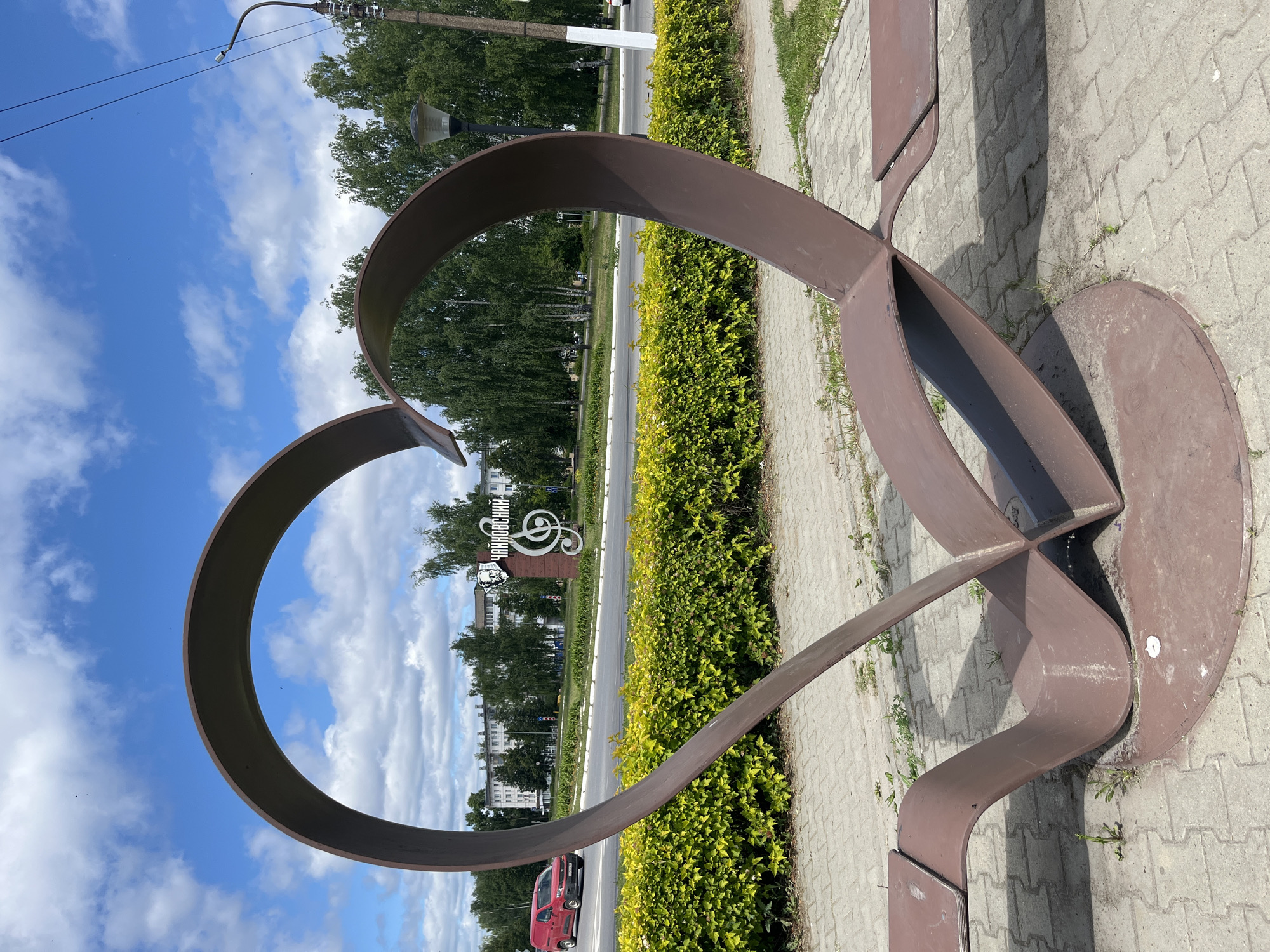 The image shows a large, brown, heart-shaped sculpture with integrated seating in the foreground, located in the city of Chaikovsky, Russia. The sculpture is positioned on a paved area with sparse grass growth. Behind the sculpture, a neatly trimmed green hedge runs along a road. Across the road, a red van is visible, and in the background, there are multi-story buildings and trees. A prominent sign with a portrait of Pyotr Ilyich Tchaikovsky and a treble clef symbol is also visible, indicating the location is likely related to the composer. The sky is blue with scattered white clouds. A lamppost stands to the right of the sculpture.