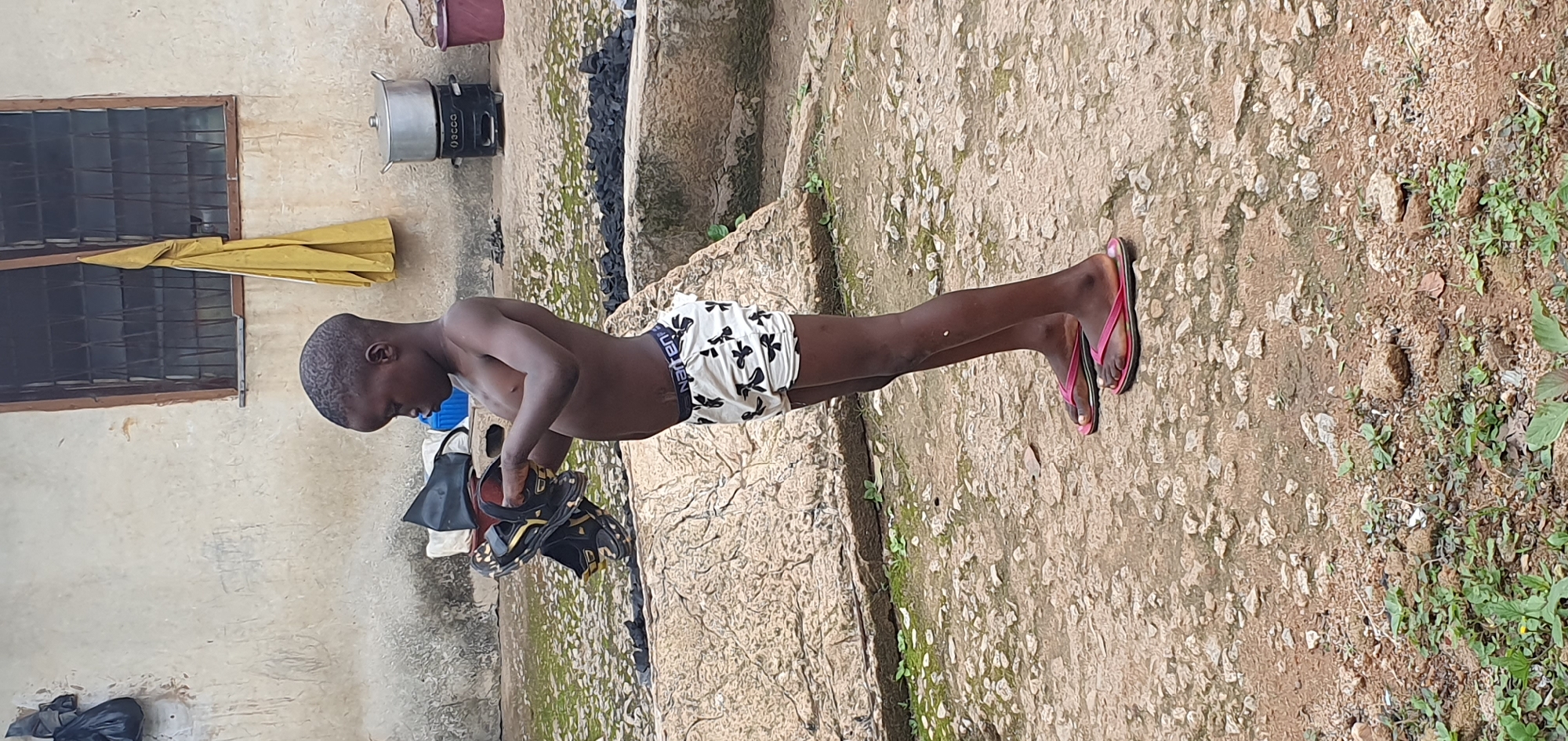 A young boy, likely under ten years old, stands outside in what appears to be a courtyard. He is shirtless, wearing patterned boxer shorts, and pink flip-flops. In his hands, he holds a pair of dark-colored sandals. The ground is made up of dirt and scattered stones. Behind the boy, a rough-textured wall with a barred window is visible. A yellow garment hangs from the window. To the right, a silver pot sits atop a black stove. In the background, a large rock formation is present. The overall scene suggests an outdoor domestic setting, possibly in a rural or less developed urban area. There is no specific indication of the city of Anguldi, Nigeria, present in the image.