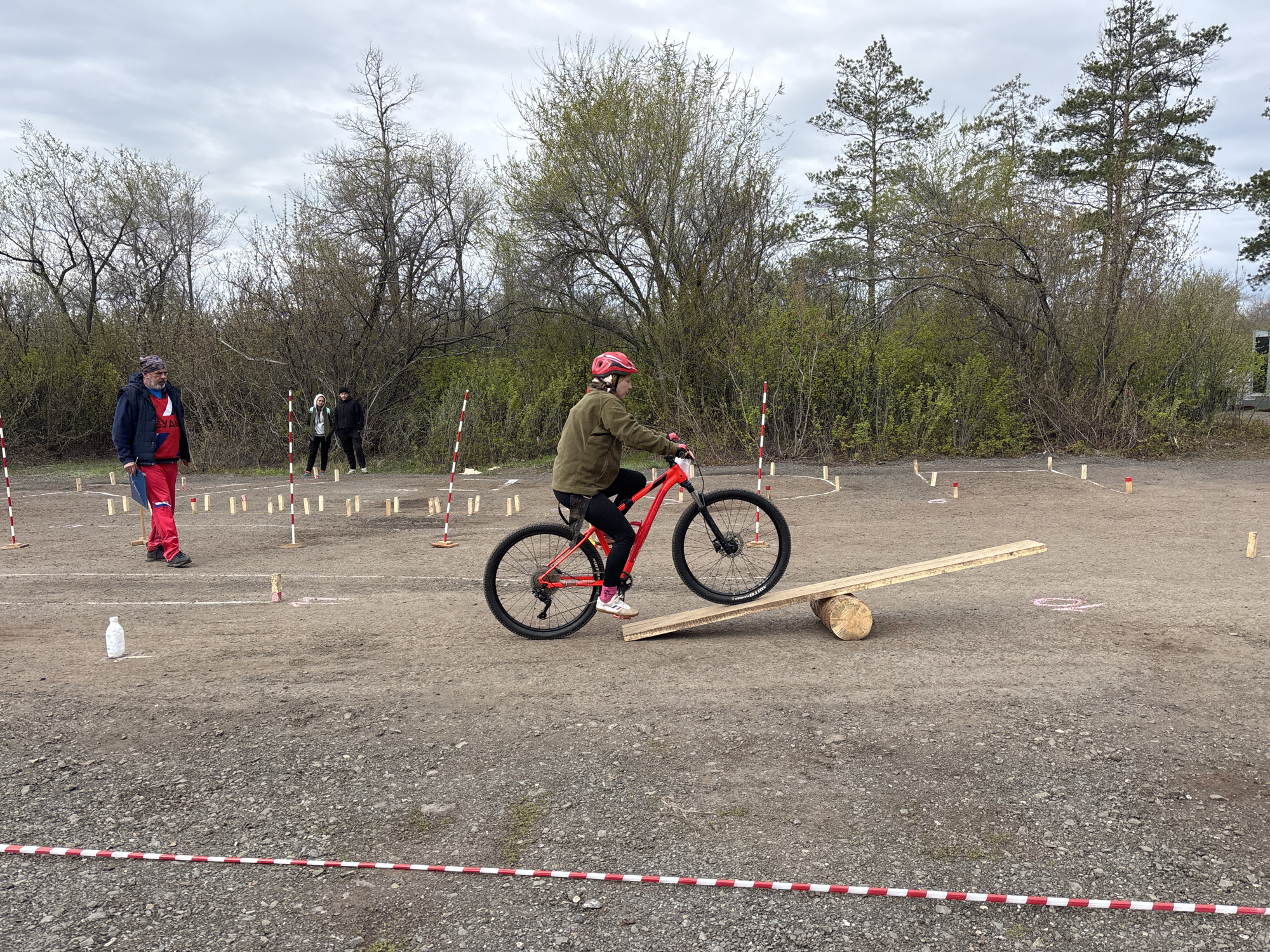 The image depicts a person riding a red bicycle on a wooden seesaw obstacle. The setting appears to be an outdoor obstacle course, marked by red and white striped boundaries and wooden poles. A person wearing a red and blue jacket stands to the left of the cyclist, possibly observing or officiating. Two other individuals are visible in the background, standing near more poles. The ground is covered in gravel. The background contains sparse trees, and the sky is overcast. The location is identified as Orenburg, Russia.