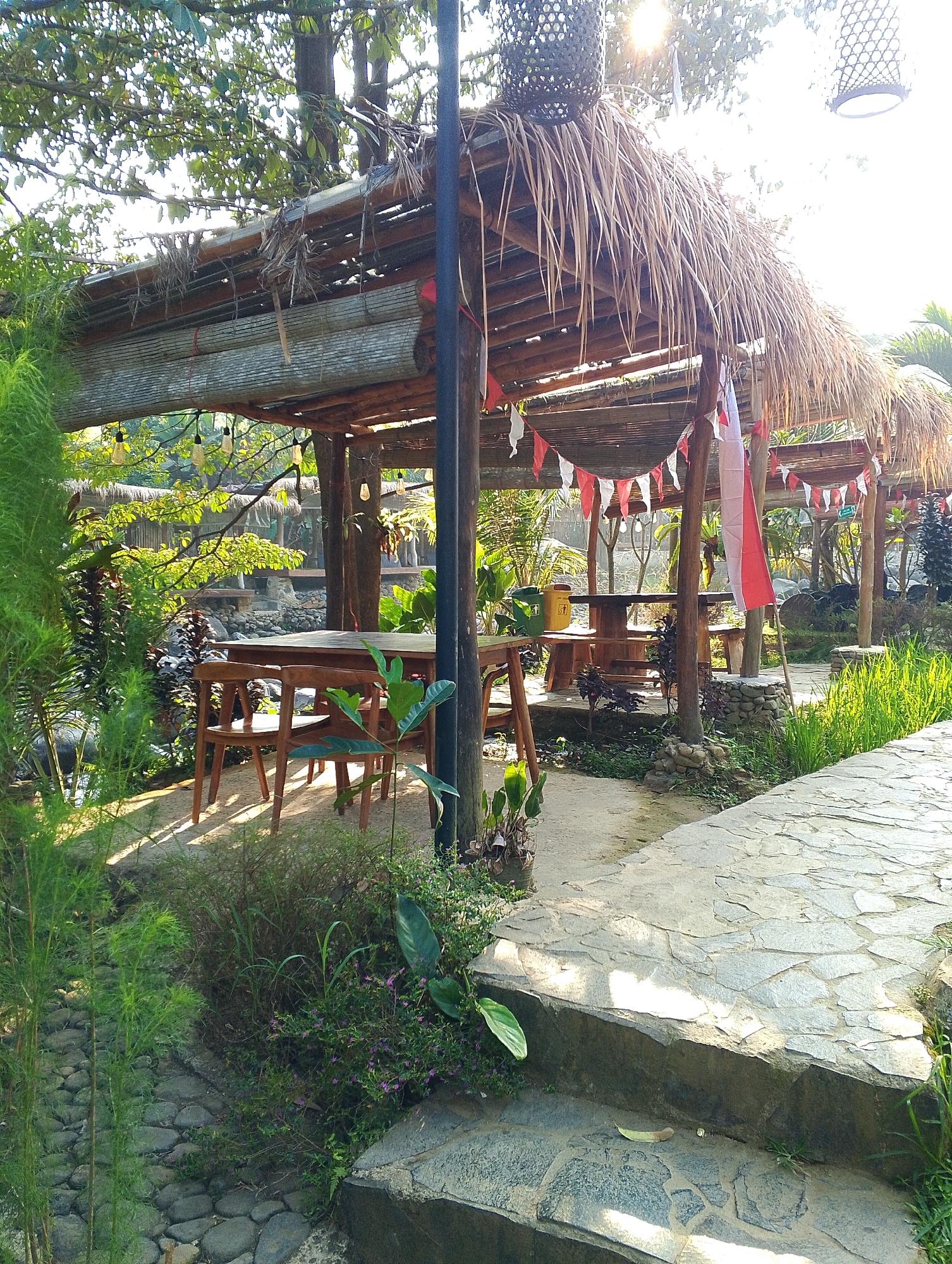 The image depicts an outdoor seating area with thatched-roof gazebos, likely a cafe or restaurant, located in Babakan Madang, Indonesia. The foreground features a stone pathway leading up a set of stairs, with lush greenery and tall, feathery plants to the left. In the midground, under one of the gazebos, a wooden table and chairs are visible, bathed in dappled sunlight. The gazebos themselves are constructed with natural materials, including bamboo and dried leaves for the roofing. Small red and white triangular flags are strung along the structures, suggesting a festive or nationalistic theme. The background reveals more of the natural environment, with trees and sunlight filtering through the leaves. The overall atmosphere is tranquil and tropical, with elements of rustic charm. No people are visible in the image.