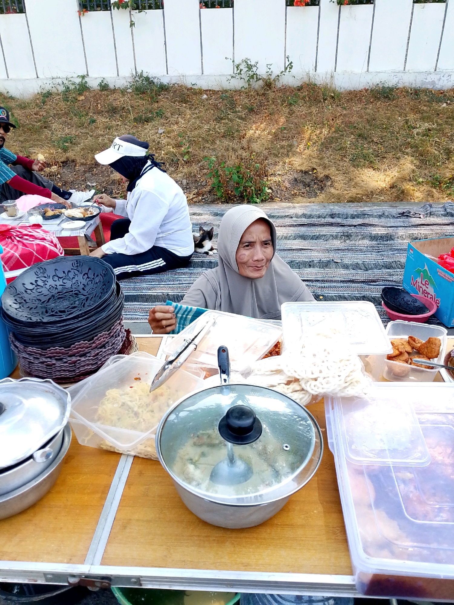 In Bulusari, Indonesia, a street food vendor in a grey hijab displays her wares from a foldable table. Behind her, a small dog with black and white markings rests on a striped mat. To her left, two people sit on the ground, one in a white long-sleeved shirt and white cap, and another in a red and blue striped shirt and dark pants, both appearing to be eating. The vendor's table is laden with food items, including fried snacks in plastic containers, a stack of black plates, and pots of food. A blue water bottle with "Le Minerale" branding is visible in the background. The environment suggests an outdoor setting, possibly a park or roadside, with dry grass and vegetation visible behind a white wall.
