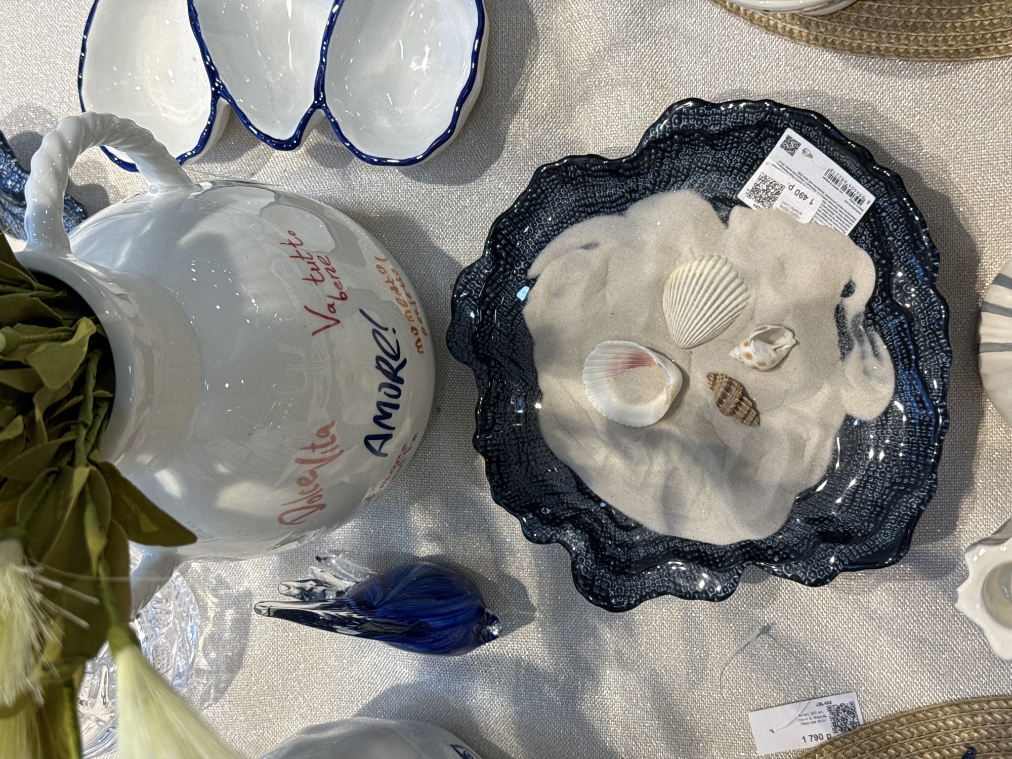 This is a close-up, overhead shot of various decorative items displayed on a textured, light-colored surface, likely in a retail setting in Krasnoyarsk, Russia.
A white ceramic pitcher with a twisted handle is adorned with Italian phrases like "Dolce Vita," "AMORE!" and "Va, tutto bene," inscribed in different colors. The pitcher appears to contain green and white artificial foliage.
Beside the pitcher is a decorative, dark blue wavy-edged dish filled with sand and several seashells. A price tag is affixed to the dish, indicating a price of 1490 rubles. A second tag in the lower right indicates a price of 1790 rubles.
A small, blue glass bird figurine rests near the pitcher. Portions of other white ceramic items, including a scalloped dish with a blue rim, are visible in the periphery of the frame.
The overall impression is a staged display in a shop selling home decor and souvenirs.