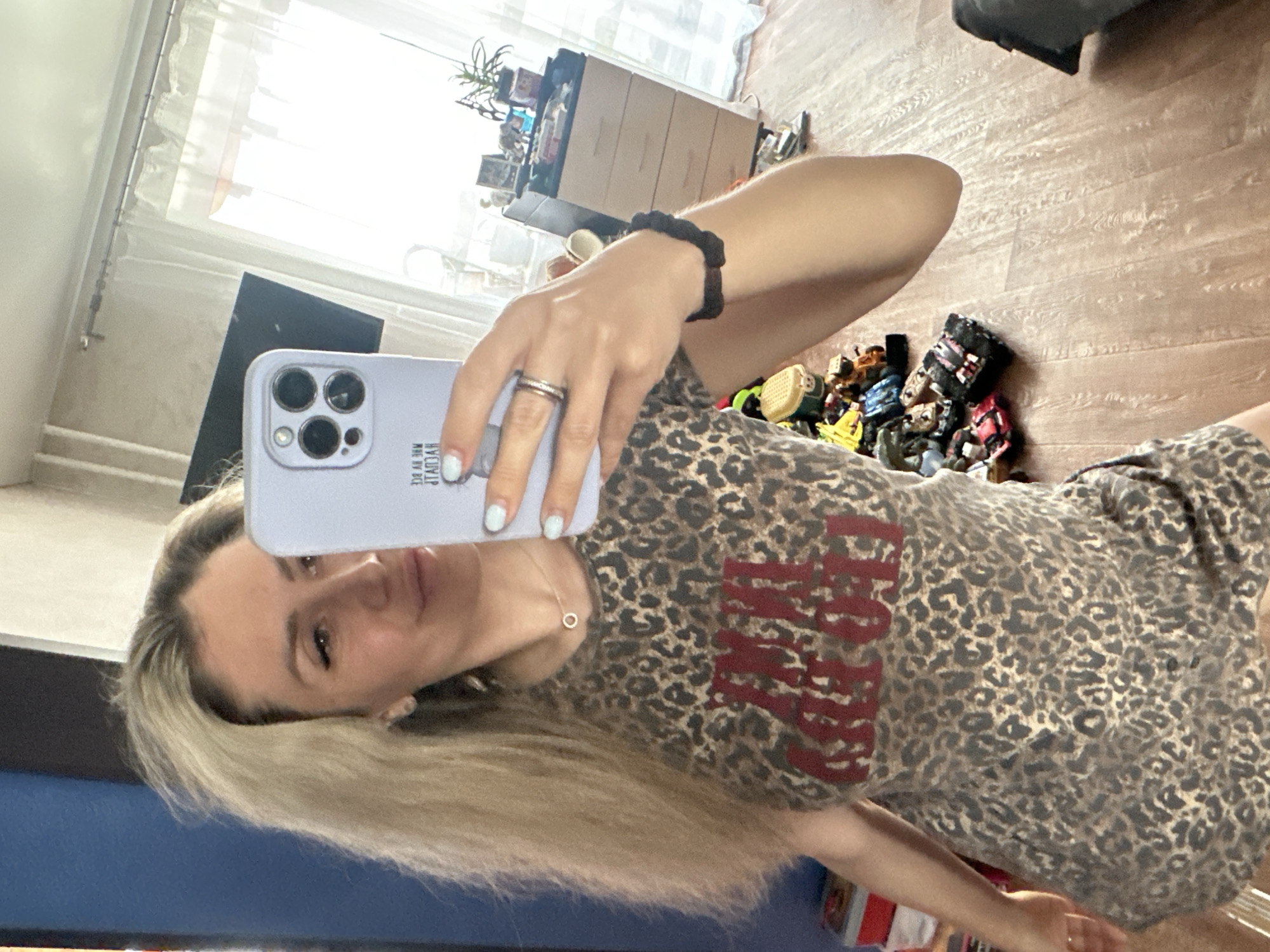 A woman is taking a selfie in a room. She has long blonde hair and is wearing a leopard-print dress with red lettering. She is holding a light purple smartphone in her right hand. The room has wooden floors and a window with white curtains. There are some toys scattered on the floor near a chest of drawers.
