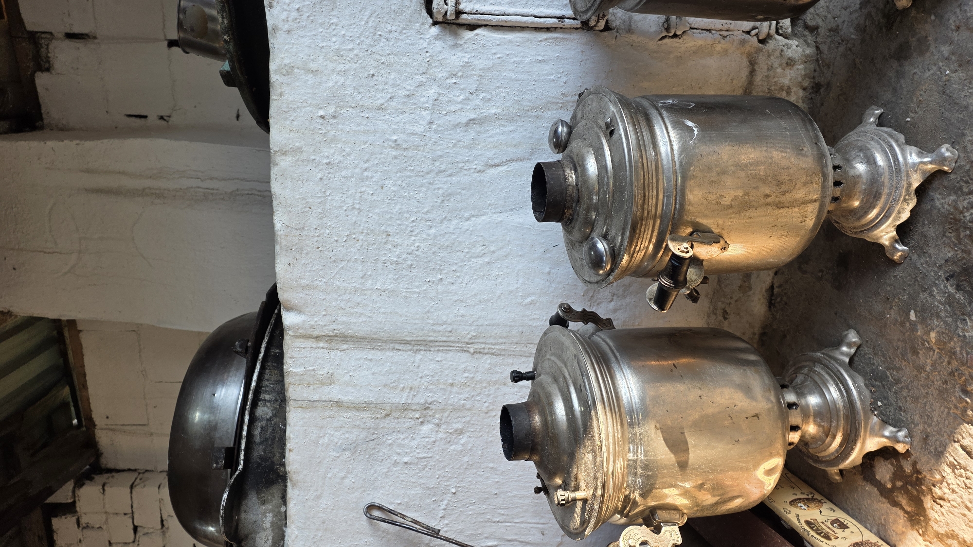 The image depicts an indoor scene, possibly a kitchen or storage area, in Terek-Suu, Kyrgyzstan. Two traditional Russian samovars, made of polished metal, stand on a rough stone floor in the foreground. Both samovars are cylindrical with ornate bases and tops, featuring handles and spouts. The samovar on the left is slightly larger and appears to be positioned in front of a decorative item with a repeating pattern. Behind the samovars, a rough, white-plastered wall serves as the backdrop. Above this wall, metal pots or lids are visible, resting on a shelf or ledge. The ceiling structure above is made of wood and corrugated metal. The overall environment appears aged and rustic, with evidence of soot or stains on the wall and a slightly uneven floor. The lighting suggests it is daytime, with sunlight casting shadows and highlighting the metallic surfaces of the samovars. No people are present in the image.