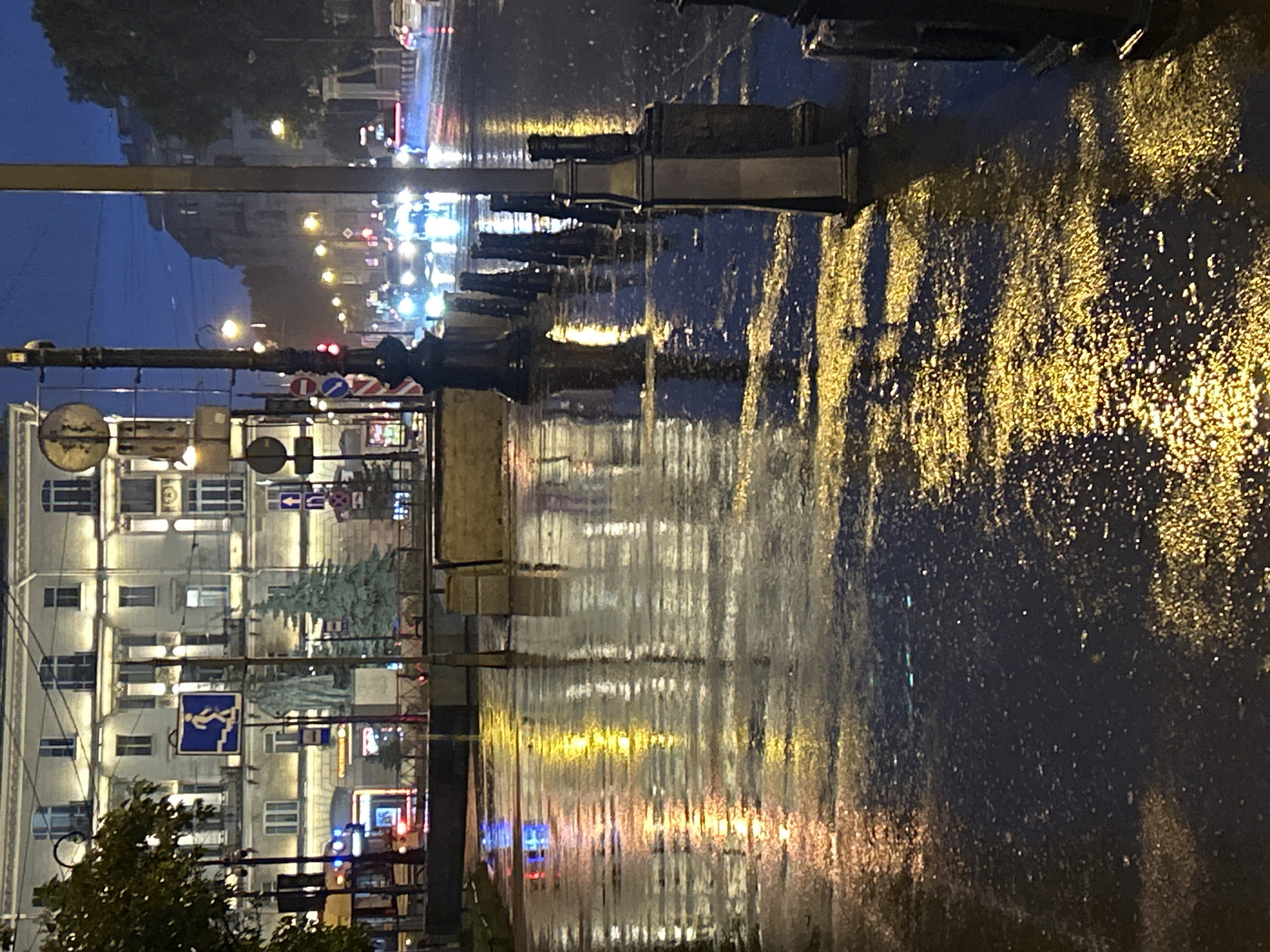The image shows a city street scene at night, likely after rain, as evidenced by the wet pavement and reflections of lights. A large, multi-story building with ornate architecture is visible on the left side of the frame. In front of the building, there is a sidewalk with bollards lining the edge. The street is wet and reflects the lights from vehicles and streetlights, creating a shimmering effect. Several cars are visible on the street, with their headlights on, suggesting they are moving. The sky is dark blue, indicating it is nighttime. There are traffic lights and street signs visible. A statue is also present in front of the building. The overall atmosphere is that of a bustling city street at night, with the wet conditions adding a moody and reflective quality to the scene.