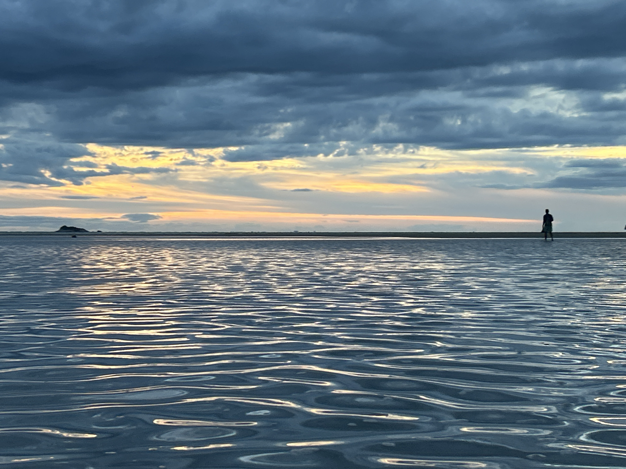 The image shows a tranquil scene at ตำบลเกาะพะงัน, Thailand. In the foreground, calm water reflects the sky above, creating a rippled pattern of light. In the midground, a flat, sandy area stretches towards the horizon. A solitary figure stands silhouetted on this sandy expanse. A small, dark landmass is visible on the left. The sky is dominated by heavy, grey storm clouds, with a band of yellow and orange light peeking through near the horizon.