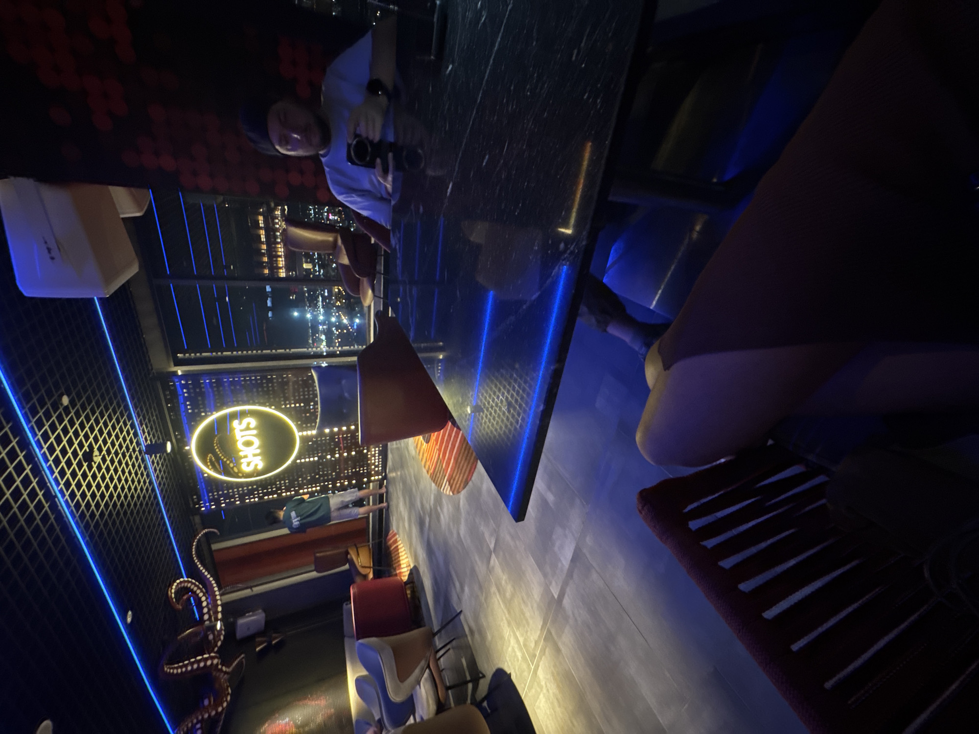 The image captures a vibrant, dimly lit bar or lounge at night, offering panoramic views of a bustling city skyline, likely in Đà Nẵng, Vietnam. The primary focus is on a person seated at a dark, reflective table, possibly made of marble or granite. This individual, wearing a light shirt and a dark cap, is holding a black camera, with their reflection clearly visible on the table surface. A brown chair with a red backrest is positioned next to the table.
In the background, through large windows, numerous city lights twinkle, indicating an urban environment from a high vantage point. Another person, dressed in a dark top and light shorts, stands further back near the window, observing the view. The interior is adorned with striking artificial lighting, including blue LED strips running along the ceiling and outlining the edges of tables, and a prominent glowing yellow neon sign that reads "SHOTS" within an oval frame.
Other notable decor elements include a large, reddish-brown octopus sculpture with white suckers, positioned on the left side of the room. Various modern chairs in different styles and colors (red, beige, blue) are scattered throughout the lounge area, suggesting a relaxed social setting. The floor features large, light grey tiles, reflecting the ambient light. The overall atmosphere is cool and sophisticated, typical of a modern city rooftop bar. The image itself is presented with a 90-degree clockwise rotation, giving an unconventional perspective of the scene.