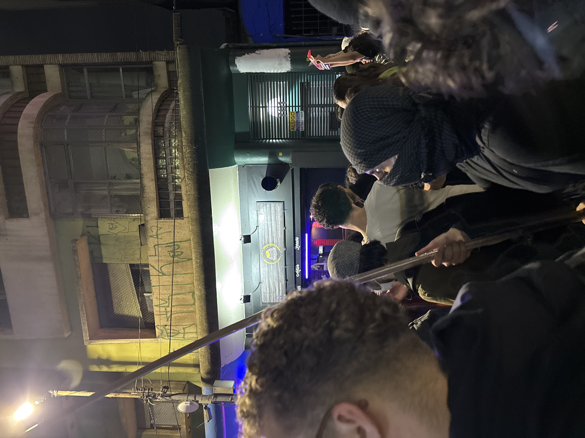 The image shows a group of people gathered in front of a building at night. The building appears to be a commercial establishment, possibly a gym or fitness center called "Urban Tribe Club," based on a sign visible on the facade. The location is most likely in the city of São Paulo, Brazil. The people are densely packed together, suggesting a social gathering, event, or demonstration. One person is holding a long, metal object, and some of the people are wearing hats. The building has a mix of architectural styles, with older sections featuring arched windows and newer additions with modern signage. There is street lighting visible, and there are some utility poles with wires nearby. The building also has an "ALLUGO" sign attached to a ventilation grill. Graffiti is also visible on the walls of the building.