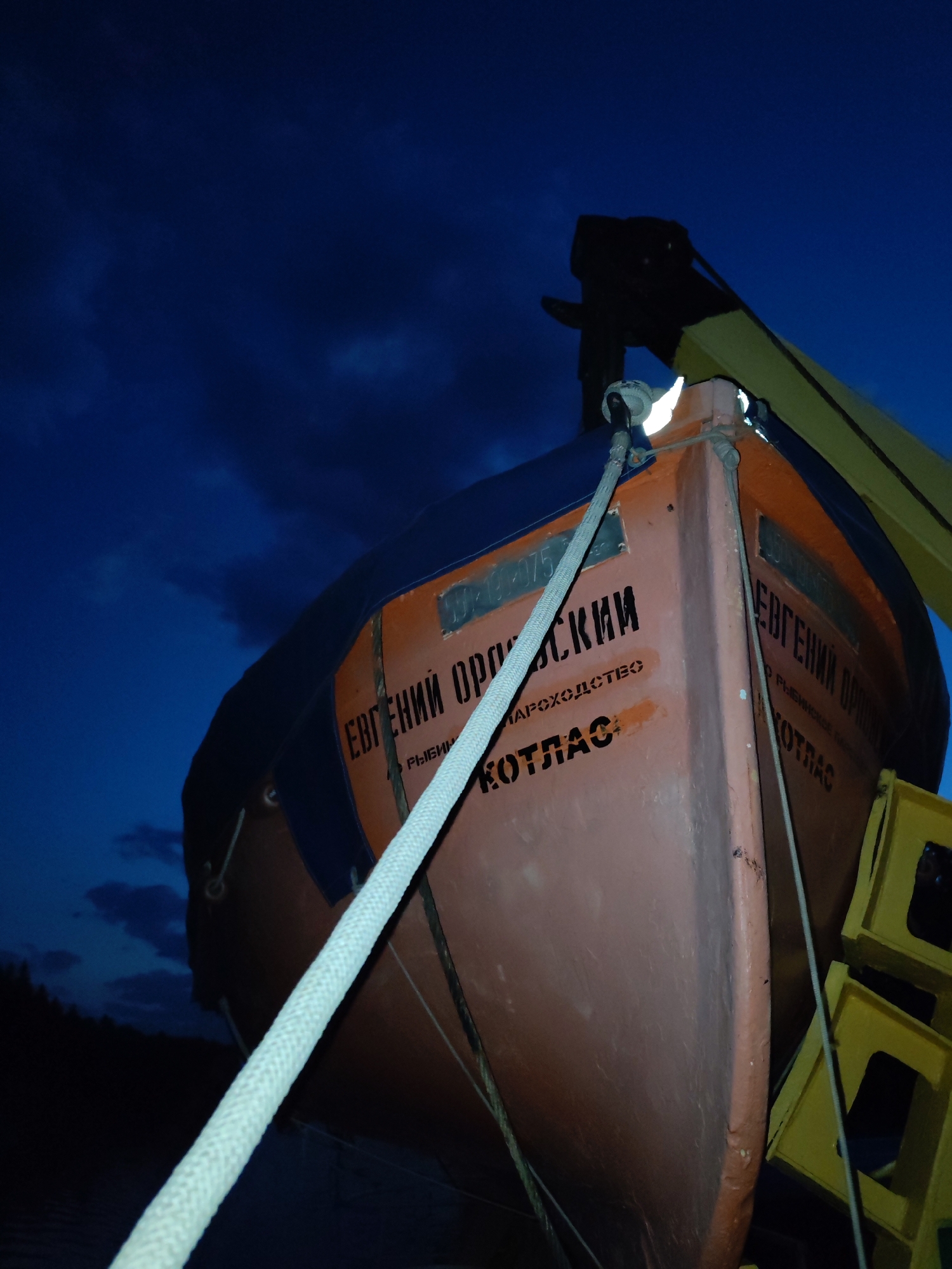 The image shows a lifeboat, labeled "Евгений Орловский" (Evgeniy Orlovsky) and "Котлас" (Kotlas), suspended from a crane or davit under a dark, cloudy sky. The location details state that the boat is located in the city of Unknown, Unknown. The lifeboat is orange with black lettering and a blue cover. A thick white rope runs from the front of the boat upward to the crane. Yellow structures support the boat on its right side.