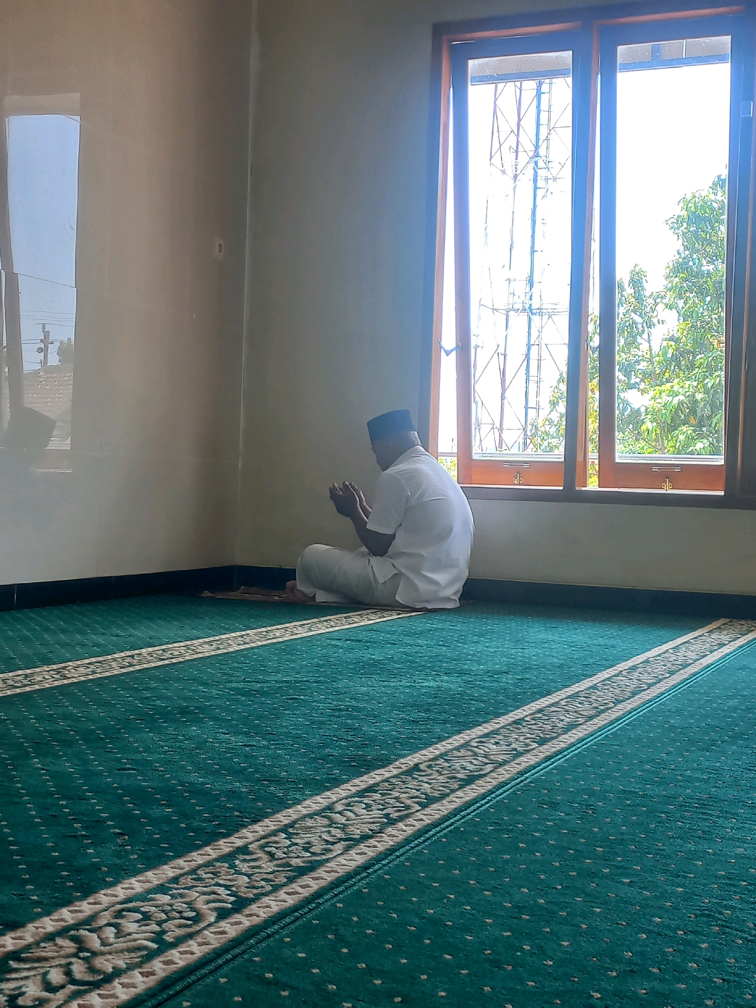 The image depicts a man wearing a white shirt and trousers, and a black cap, kneeling on a prayer mat on the floor of a room. He is facing a large window and has his hands clasped in front of him, suggesting he is in prayer. The room has plain light-colored walls and a dark skirting board. The floor is covered with a green carpet featuring a patterned border. Beyond the window, a radio tower structure is visible against a bright sky, along with green trees. The scene appears to be inside a building, possibly a mosque or a prayer room, located in Nguntoronadi, Indonesia, given the context provided. The lighting suggests it is daytime.