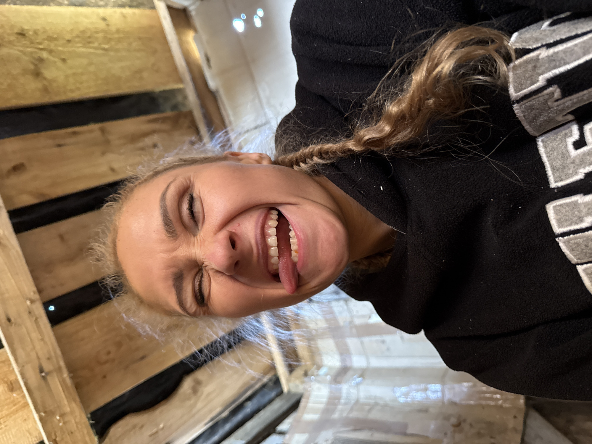 A young woman is centered, photographed up close, making a face and sticking out her tongue. She has blonde hair, with a portion of it braided and pulled back. She is wearing a black sweatshirt with white lettering visible on the right. The background appears to be a construction site or unfinished interior, with wooden planks and a white, reflective surface visible. There is no indication of Nizhnyaya Chermoda, Russia, in the image.