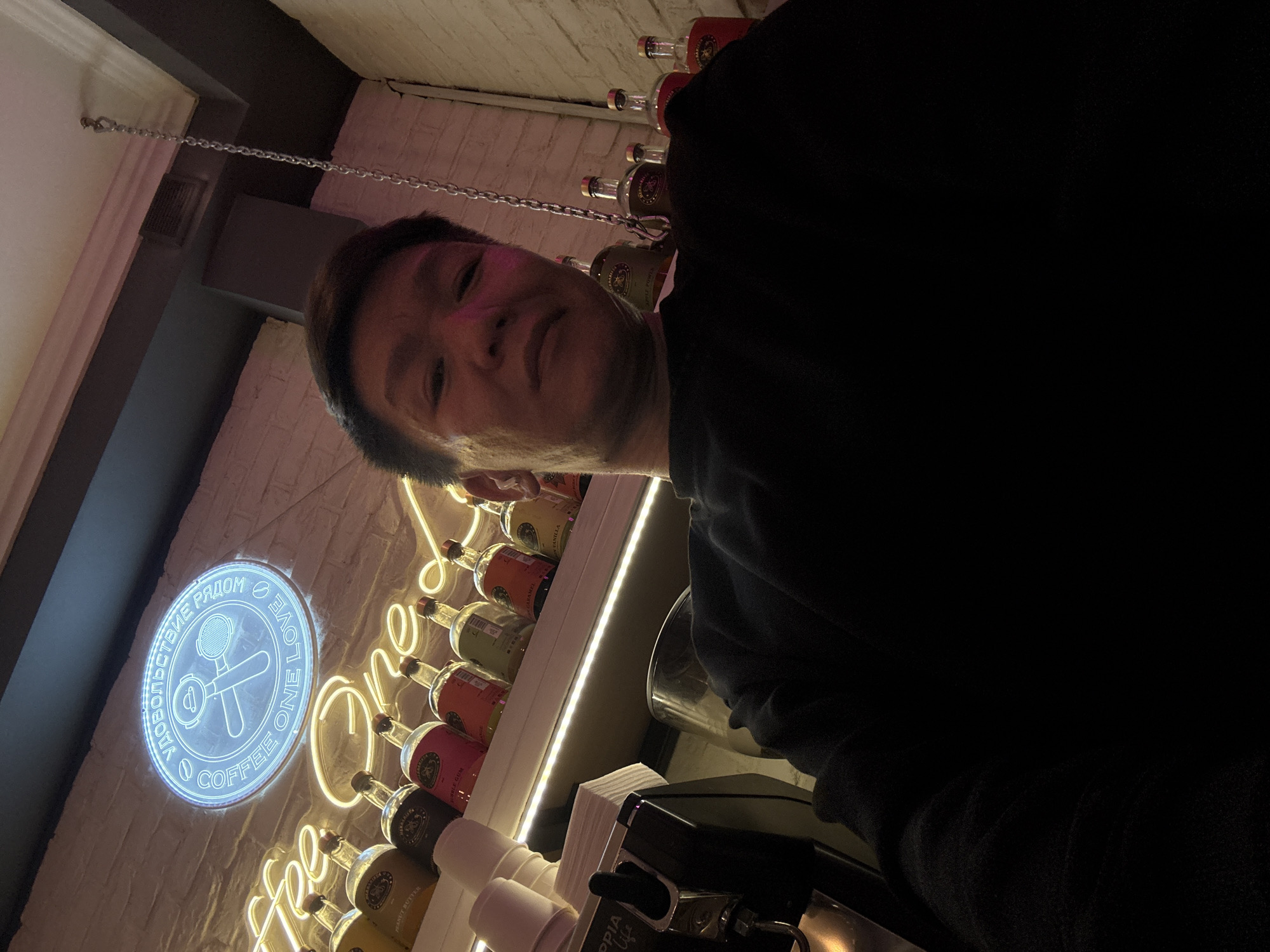 A man is taking a selfie in what appears to be a cafe or bar in Moscow, Russia. The background features a neon sign shaped like a coffee portafilter and cup, with text that includes "COFFEE ONE LOVE" and Russian characters. Shelves behind the man are lined with bottles, possibly coffee syrups or alcohol. A coffee machine is visible to the left. The lighting is dim and atmospheric, with pink and blue neon lights illuminating the space. The man is wearing a dark sweatshirt.