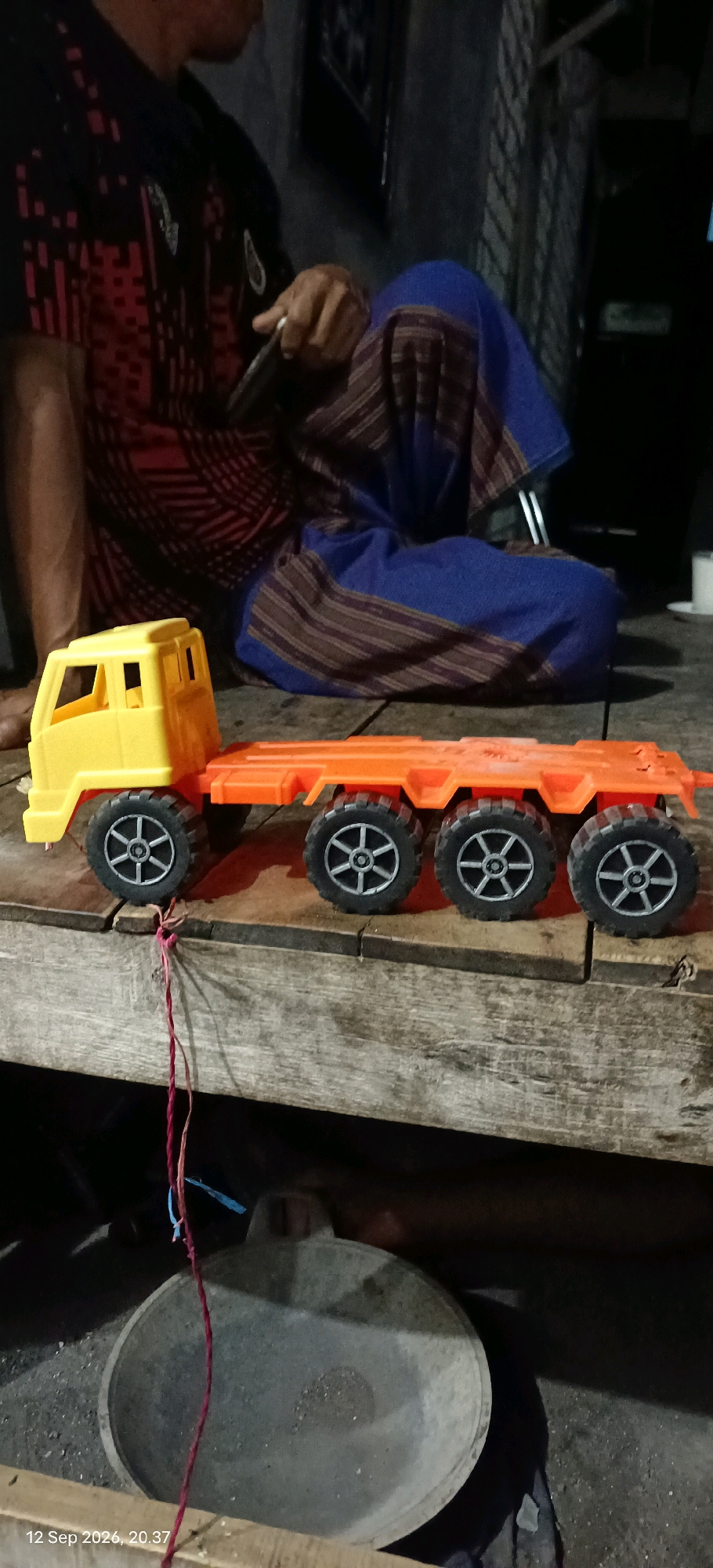In Mojopetung, Indonesia, a yellow toy truck with an orange flatbed trailer is positioned on a weathered wooden surface. The truck has six wheels visible on the trailer, each with a silver rim. Attached to the trailer by a thin red and blue string is a dark-colored pan or basin. In the background, a person is seated, partially visible, wearing a red and black patterned shirt and a blue garment with brown stripes. The environment appears to be an outdoor or semi-outdoor setting with subdued lighting.