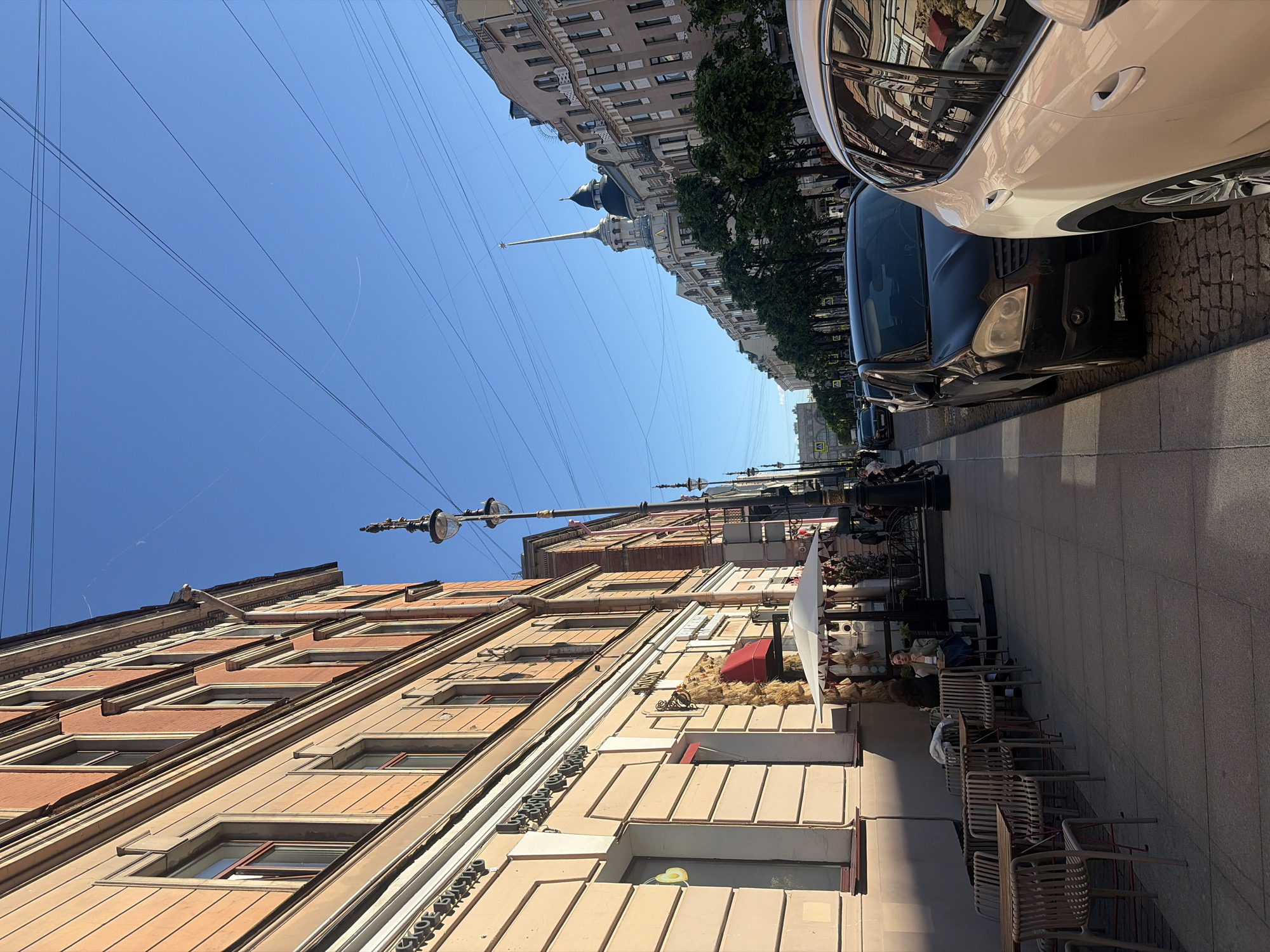 A sunny day scene on Nevsky Prospekt in Saint Petersburg, Russia. The view is from the sidewalk looking down the street, with a building on the left and cars parked along the right. The building on the left has a light orange facade with multiple windows and decorative architectural elements. In front of the building, there are tables and chairs set up outside what appears to be a cafe, with a white umbrella providing shade. Several people are seated at the tables. On the right side of the street, cars are parked in a row. The sky is clear and blue, with overhead wires visible. In the distance, the distinctive spire of the Admiralty Building can be seen.
