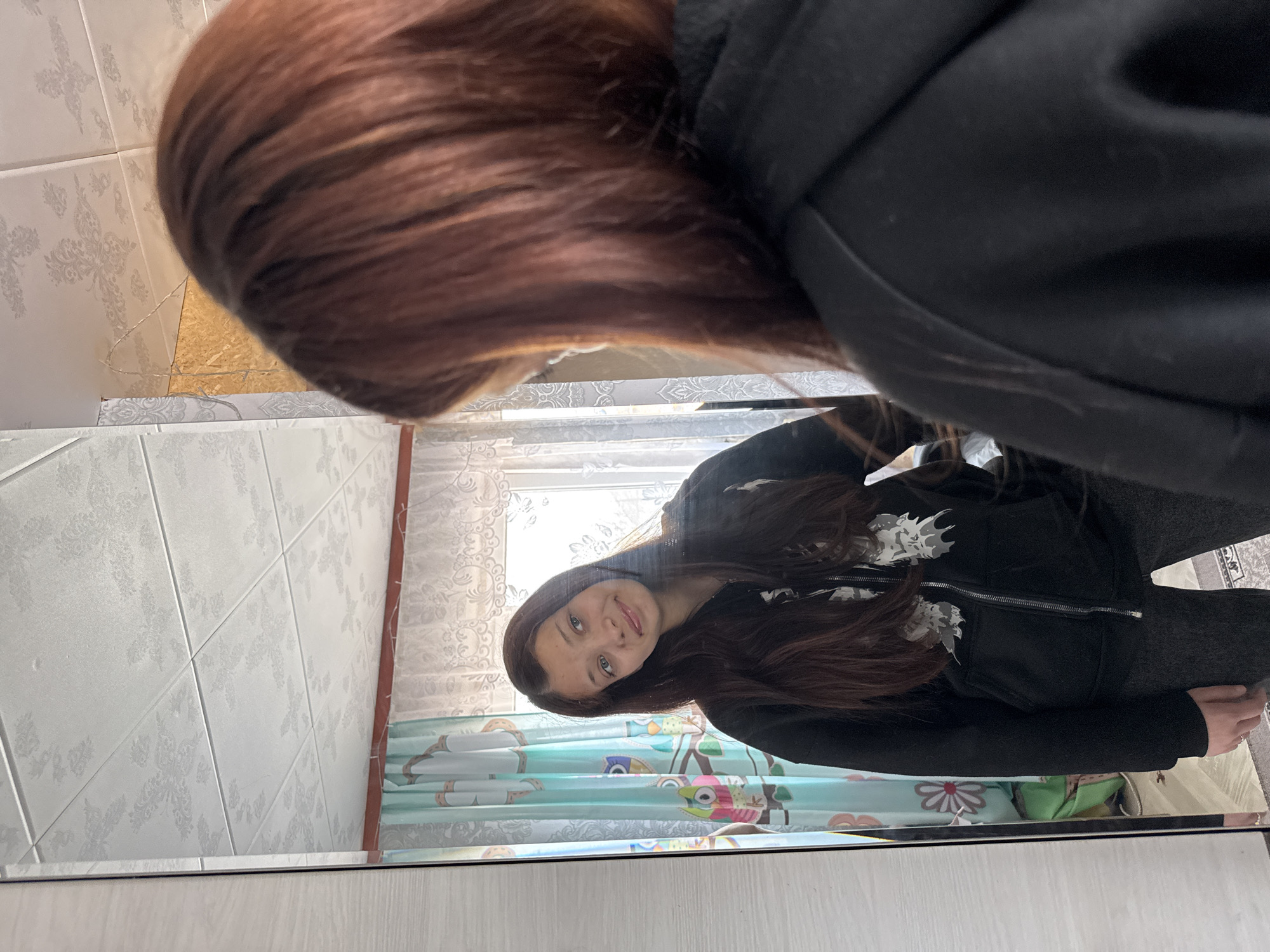 A young woman is reflected in a mirror. She is wearing a black zip-up hoodie with a white design on the chest and dark pants. Her long, dark brown hair is partially obscuring the top of the mirror. The mirror's background shows a tiled wall with a floral pattern and a window with patterned curtains. The overall scene suggests a domestic interior setting, possibly a bathroom or bedroom. The location is Кемуль, Russia.