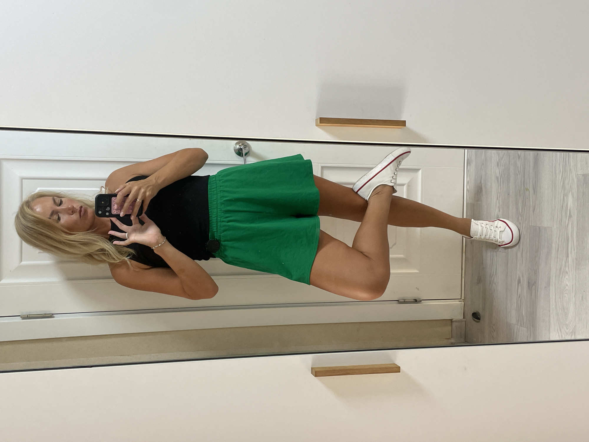 A blonde woman, seen from the knees up, stands on one leg, reflecting in a mirror. She is wearing a black tank top and bright green shorts. Her hair is shoulder-length and styled casually. She holds a smartphone in her right hand, angled as if taking a selfie. Her left leg is bent and crossed over her right knee. She is wearing white sneakers with red trim. The background shows a white door with a silver doorknob and a wooden handle on what appears to be a closet or cabinet. The floor is a light-colored wood-grain laminate. The scene appears to be indoors, likely in a residential setting. There is no visible indication of the city of Baranovka, Russia, or any other specific location.