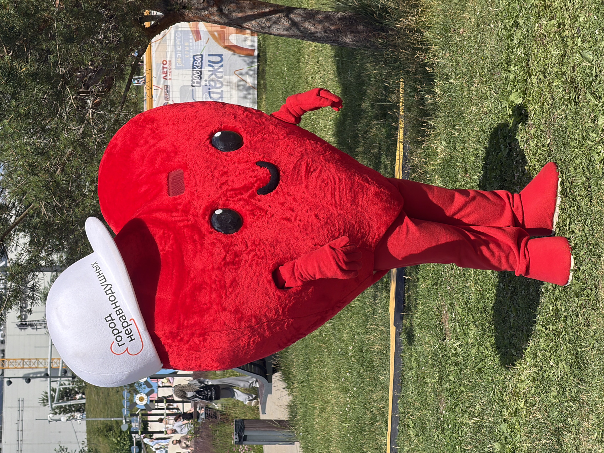 A person in a red heart-shaped costume with a white baseball cap stands on a grassy area. The cap has red text and a heart symbol. The costume has cartoonish eyes and a smile. In the background, there is a paved walkway, a bench, a trash can, some trees, and buildings. A crane is visible in the distance. There are other people in the background, some walking and some sitting. A banner with Russian text and images is visible behind the heart costume. The scene appears to be outdoors in a public area.