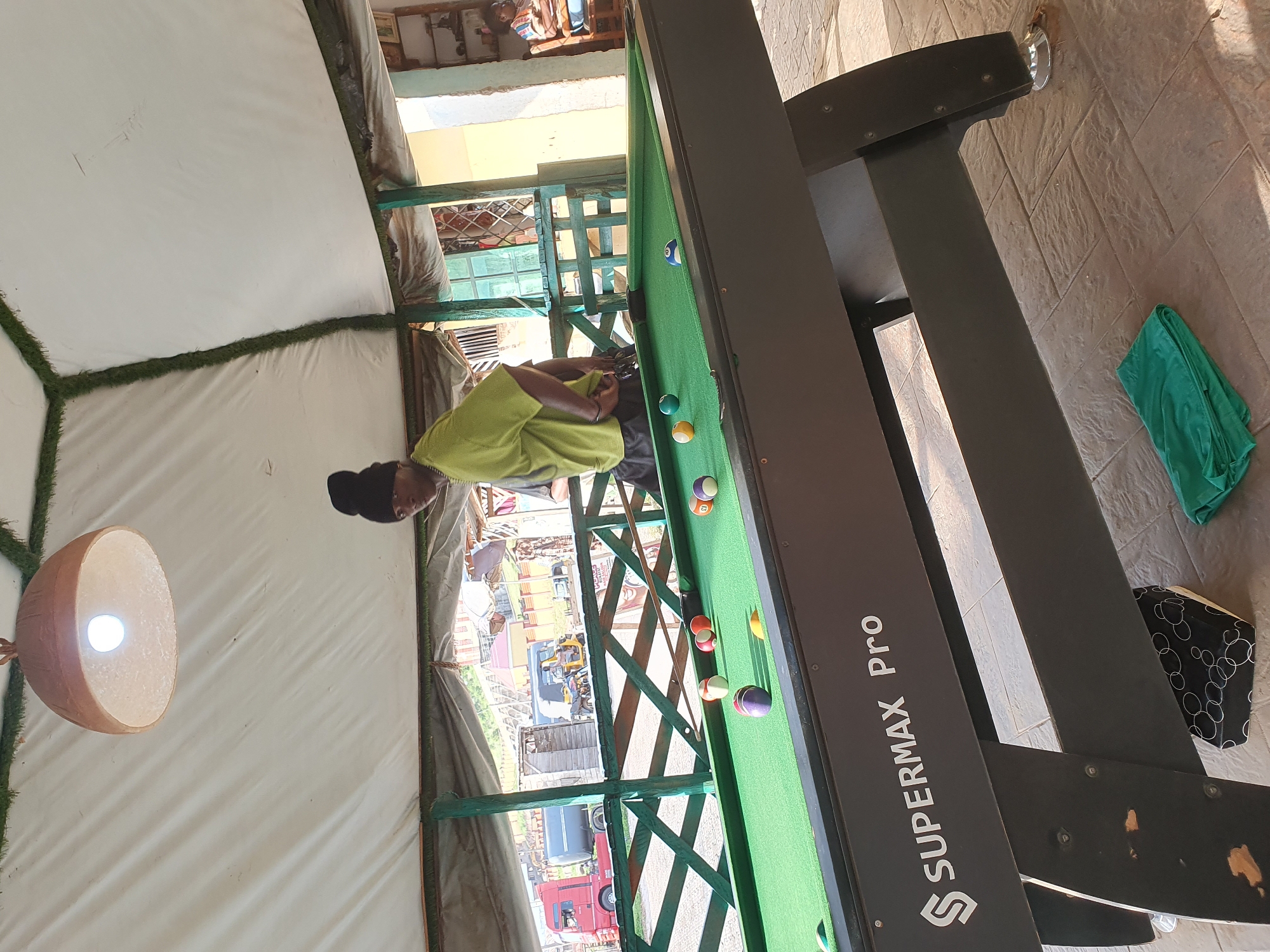 In Lokoja, Nigeria, a person wearing a black beanie and a lime green t-shirt stands near a pool table, preparing to play. The pool table, identified as a "SUPERMAX Pro," is covered in green felt and has several colored billiard balls scattered across it, along with a cue stick. The scene is outdoors, under a white tent-like structure adorned with fake grass along its edges. In the background, a red truck, a dark-colored tank truck, and a yellow motorized tricycle are visible, suggesting a street or roadside location. The environment appears to be a casual outdoor recreational area.