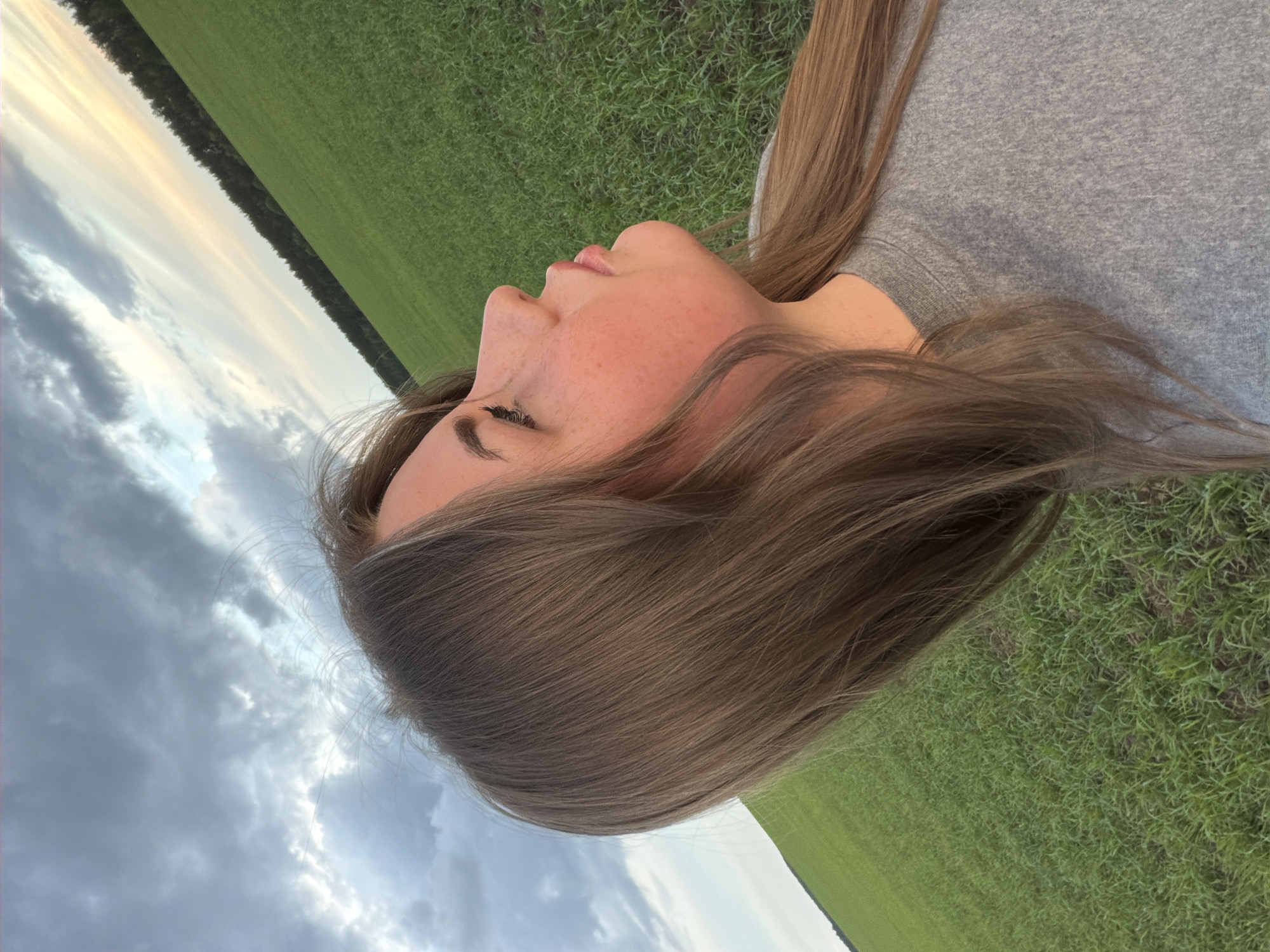 A woman lies on her side in a grassy field under a cloudy sky. She is fair-skinned with freckles and light brown hair. She wears a gray t-shirt. The field extends to a tree line. The sky is visible in the background with patches of blue and gray clouds. The location may or may not be Фоки, Russia; the image provides no visual confirmation of the location.
