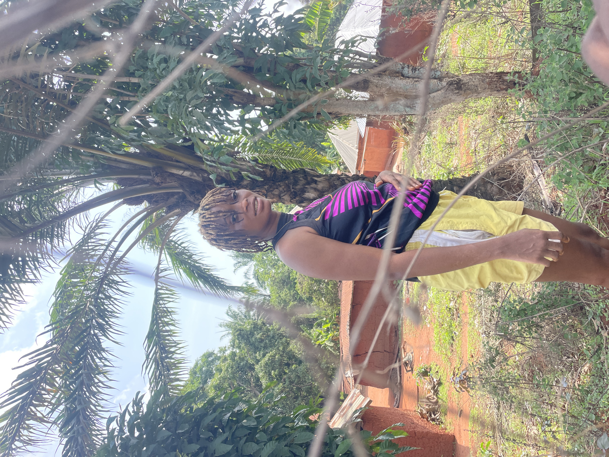The image, originally rotated ninety degrees clockwise, features a young woman posing outdoors in a bright, natural environment. She stands slightly off-center, wearing a black sleeveless top with pink and purple geometric patterns and bright yellow shorts. Her hair is styled in dreadlocks or braids, and she looks directly at the camera with a pleasant, subtle smile. Her left arm is slightly bent, and her right hand rests near her hip.
The foreground is partially obscured by several thin, out-of-focus branches and twigs, creating a layered visual effect. In the background, a prominent palm tree with large fronds dominates the left side. The entire background is filled with lush green foliage from various trees and plants. To the right of the woman, a rustic building with reddish-brown walls, likely made of mud or brick, and a corrugated metal roof is visible. The ground is a mix of reddish soil and patches of grass and weeds. Patches of clear blue sky are visible through the tree canopy, indicating bright daytime conditions. There is no visible text in the image. The setting suggests a rural area, consistent with the provided location context of Ogbe, Nigeria.