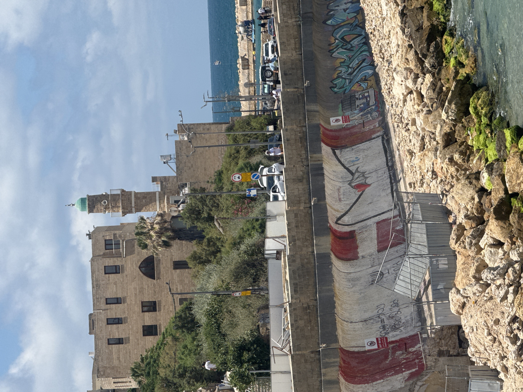 The image depicts the old city of Jaffa in Tel-Aviv, Israel, showcasing a historical building with a minaret in the background. In the foreground is a sea wall adorned with graffiti, facing the Mediterranean Sea. A road runs parallel to the wall, with parked cars and pedestrians visible. Vegetation is interspersed between the buildings and along the coastline. Moss grows on the rocks alongside the water. Scaffolding is visible along the lower portion of the wall.
