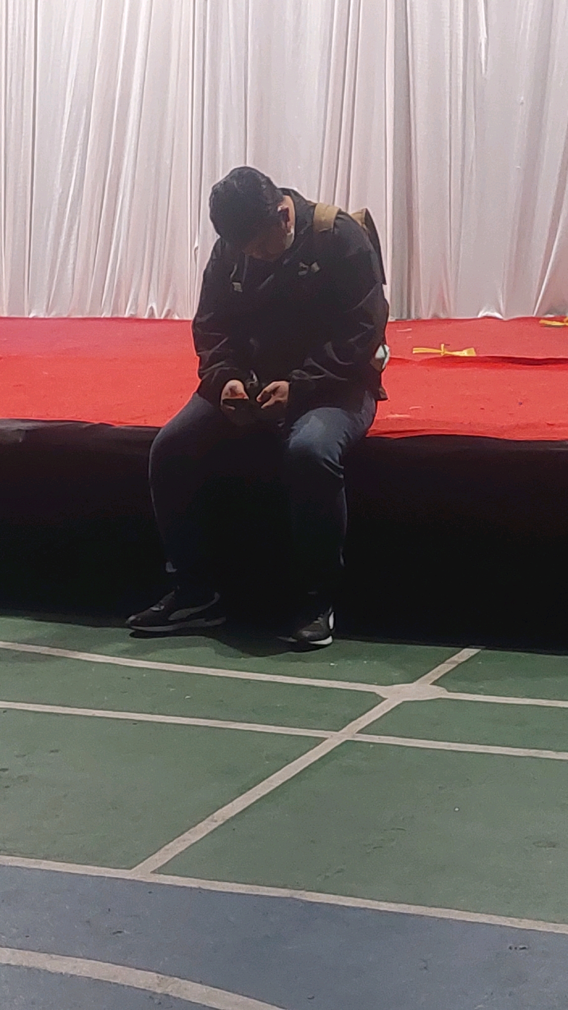 A person, appearing to be male, is sitting on a red stage or platform with a dark border. The person is wearing a dark jacket over a light-colored shirt, blue jeans, and dark athletic shoes. A backpack is visible on the person's back. The person is looking down, seemingly at something in their hands. Behind the person, a white curtain or backdrop is visible. The area in front of the stage appears to be a green court with white lines, possibly a sports court. The scene is indoors, under artificial lighting, with a slightly muted color palette. The overall impression is of a person waiting or resting in a public indoor space. Given the context of Cibinong, Indonesia, this could be a school gymnasium, a community center stage, or an indoor sports facility.