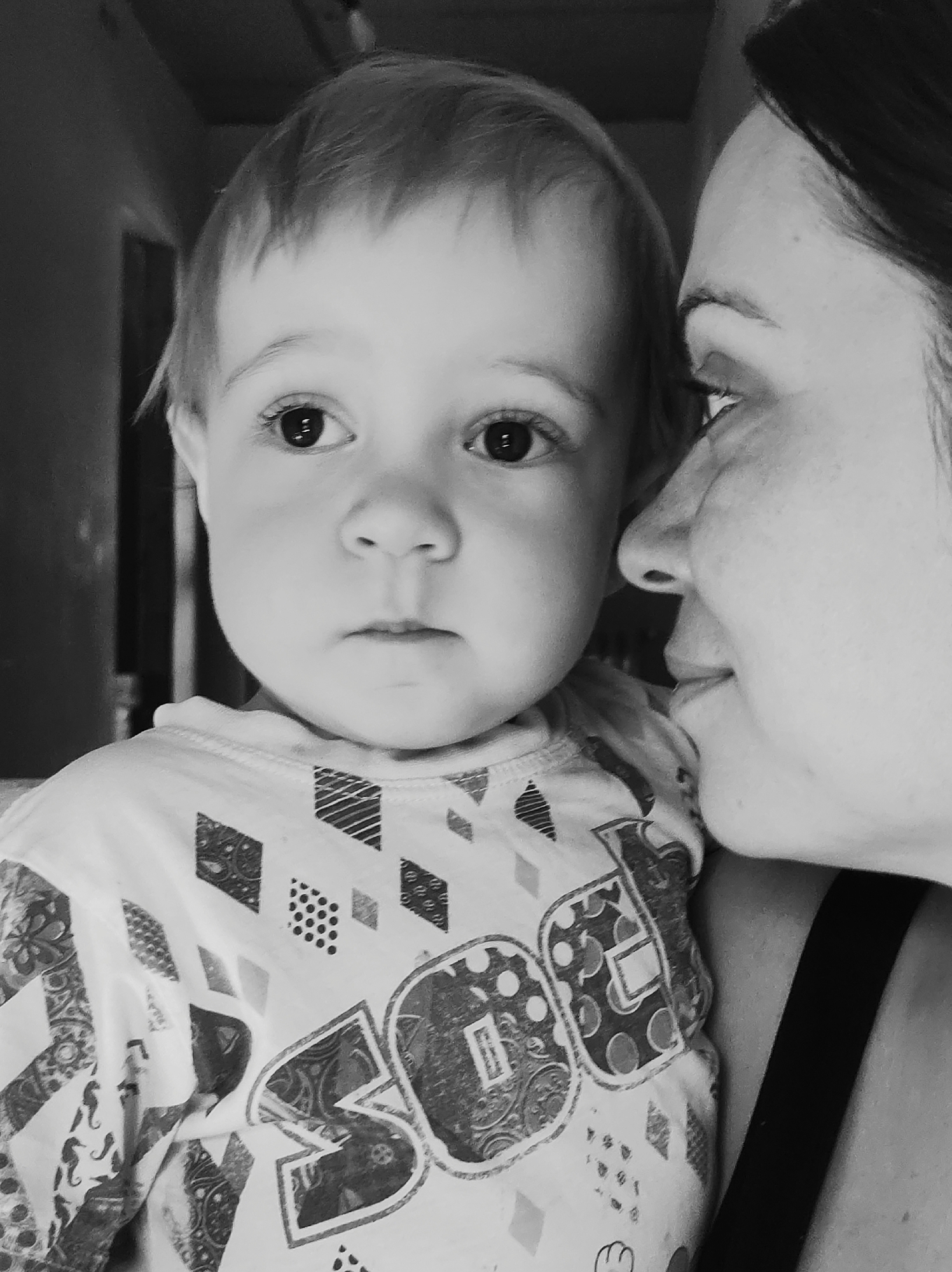A monochrome, close-up photograph captures a woman holding a young child. The child, positioned on the left, has light-colored hair, round cheeks, and dark eyes. The child wears a patterned shirt, featuring geometric shapes and the large letters "SO." The woman is positioned on the right, her face partially visible in profile. She has dark hair pulled back from her face. The background is indistinct, suggesting an indoor setting. The location Фоки, Russia, is not discernible from the visual details.